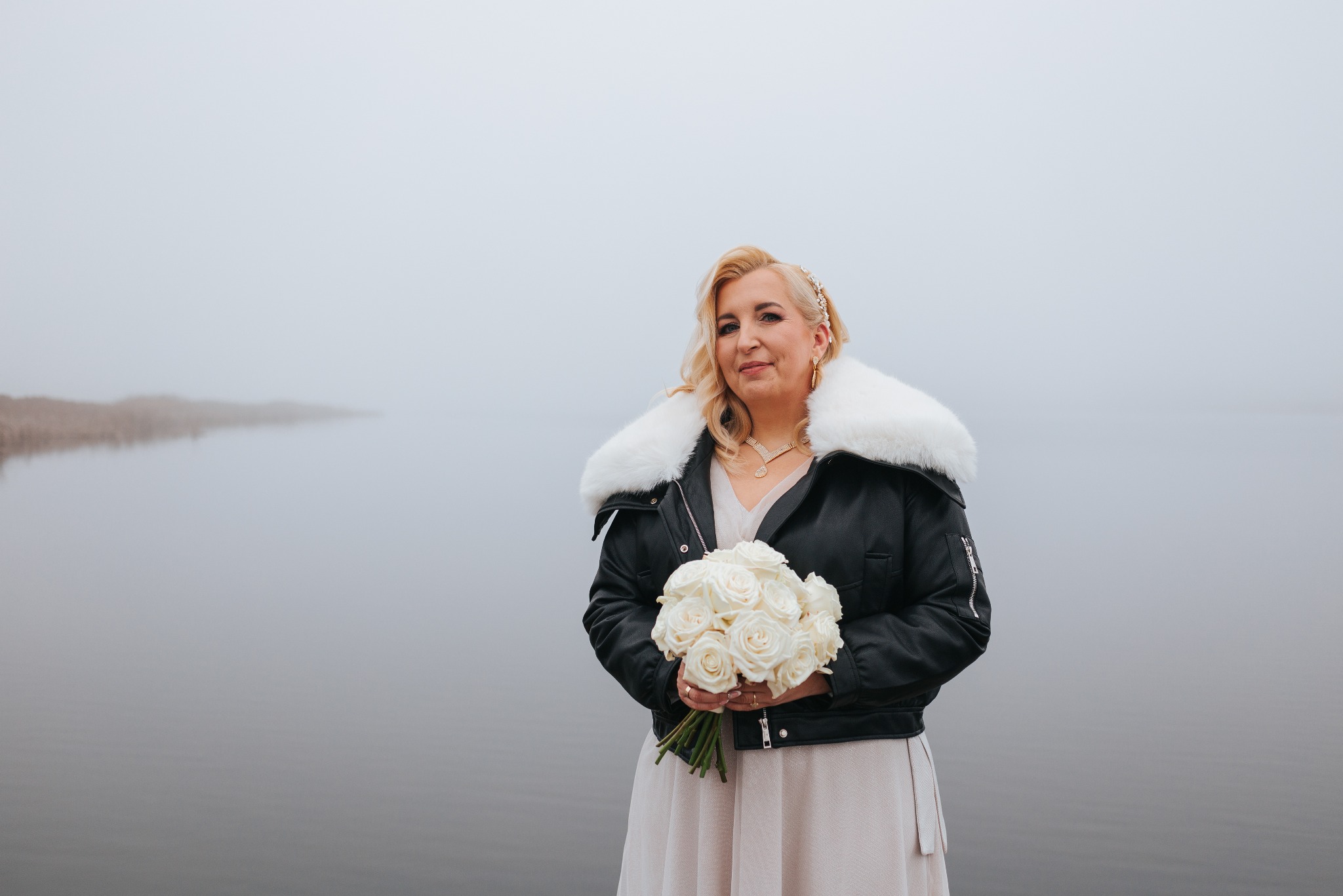 Panna młoda w skórzanej kurtce z futrzanym kołnierzem trzyma bukiet białych róż na tle mglistego jeziora. Nietypowa stylizacja ślubna.