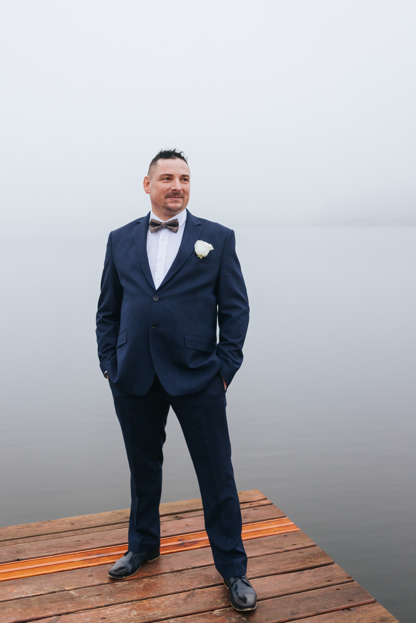 Elegancki mężczyzna w granatowym garniturze i muszce, z butonierką, stoi na drewnianym pomoście nad wodą w mglisty dzień, spoglądając w dal.