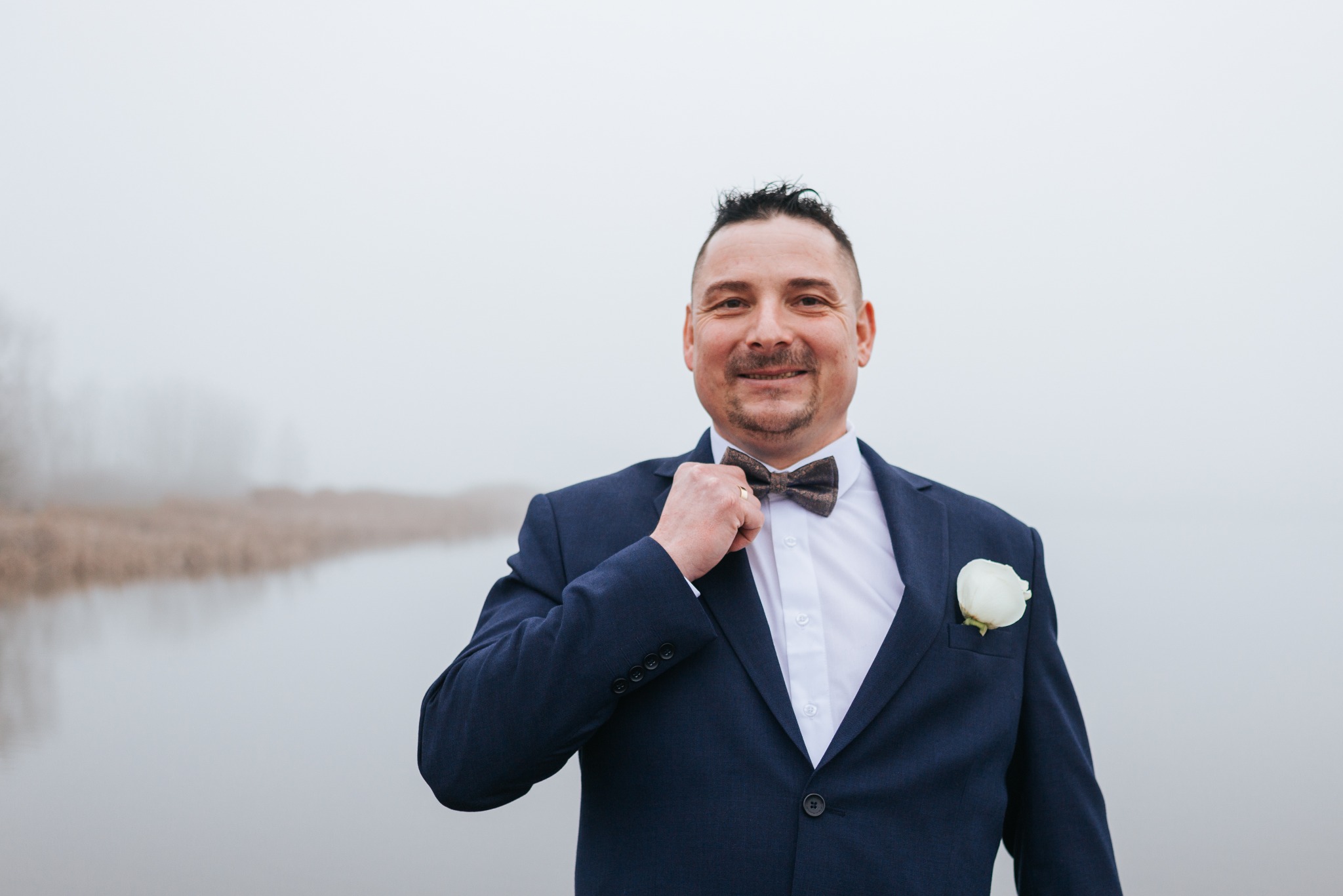 Uśmiechnięty mężczyzna w granatowym garniturze z butonierką i muszką, poprawiający ją na tle mglistego krajobrazu.