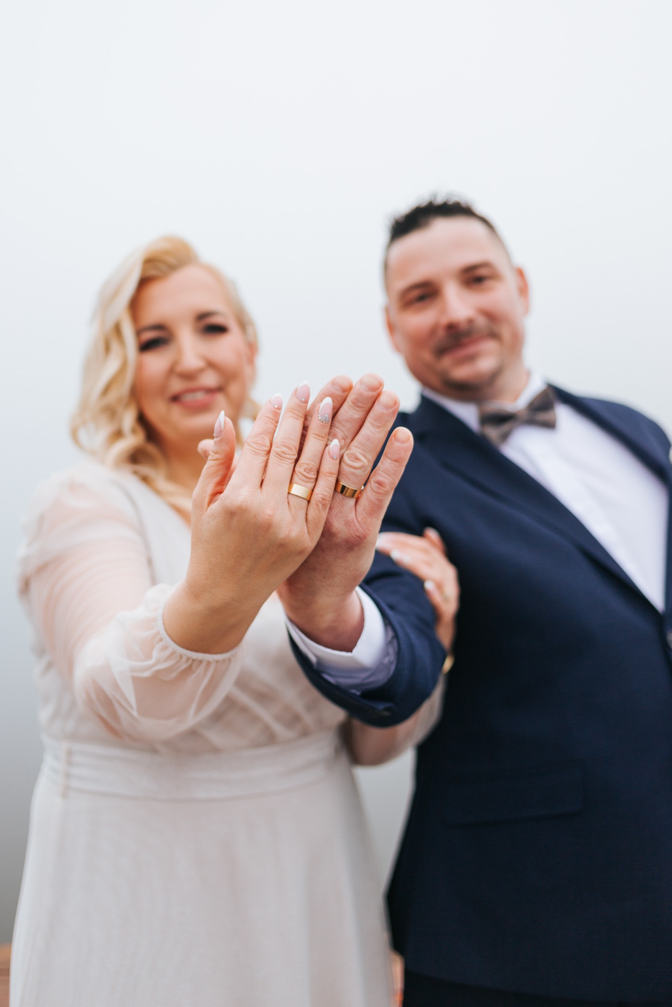 Para młoda prezentuje obrączki na dłoniach, on w granatowym garniturze z muchą, ona w białej sukni. Uśmiechnięci, w tle rozmyte tło.