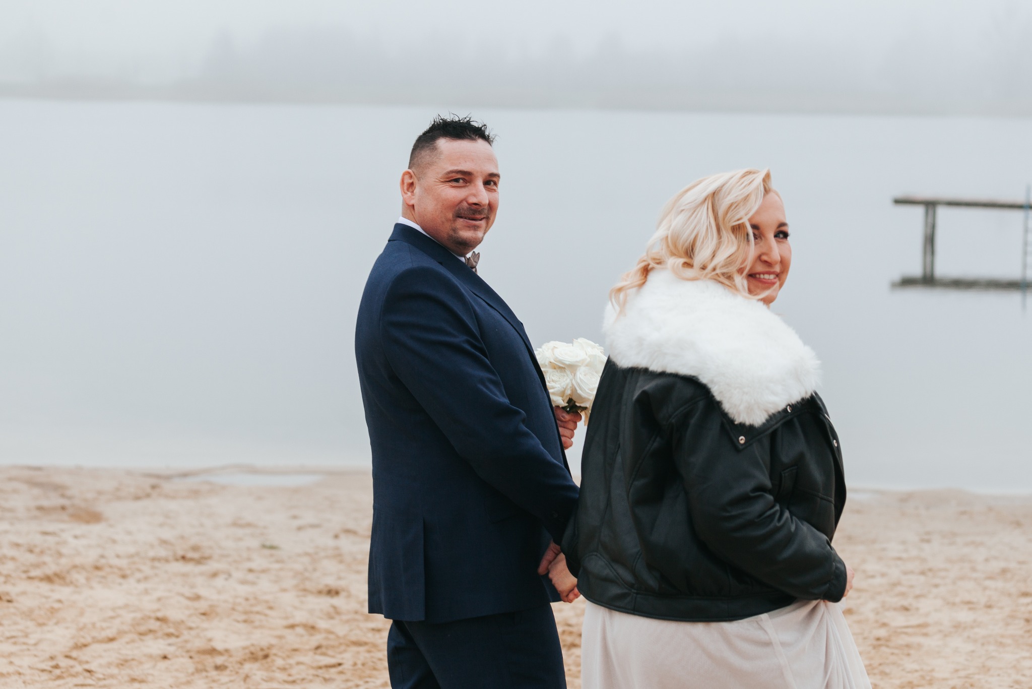 Para młoda na plaży w pochmurny dzień. Pan młody w granatowym garniturze trzyma bukiet białych róż, panna młoda w czarnej kurtce z futrzanym kołnierzem.