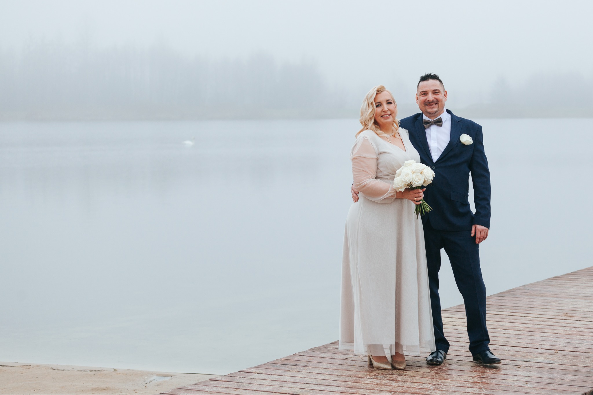 Para młoda na drewnianym pomoście nad jeziorem we mgle. Panna młoda trzyma bukiet białych róż. Pan młody w granatowym garniturze z muszką.