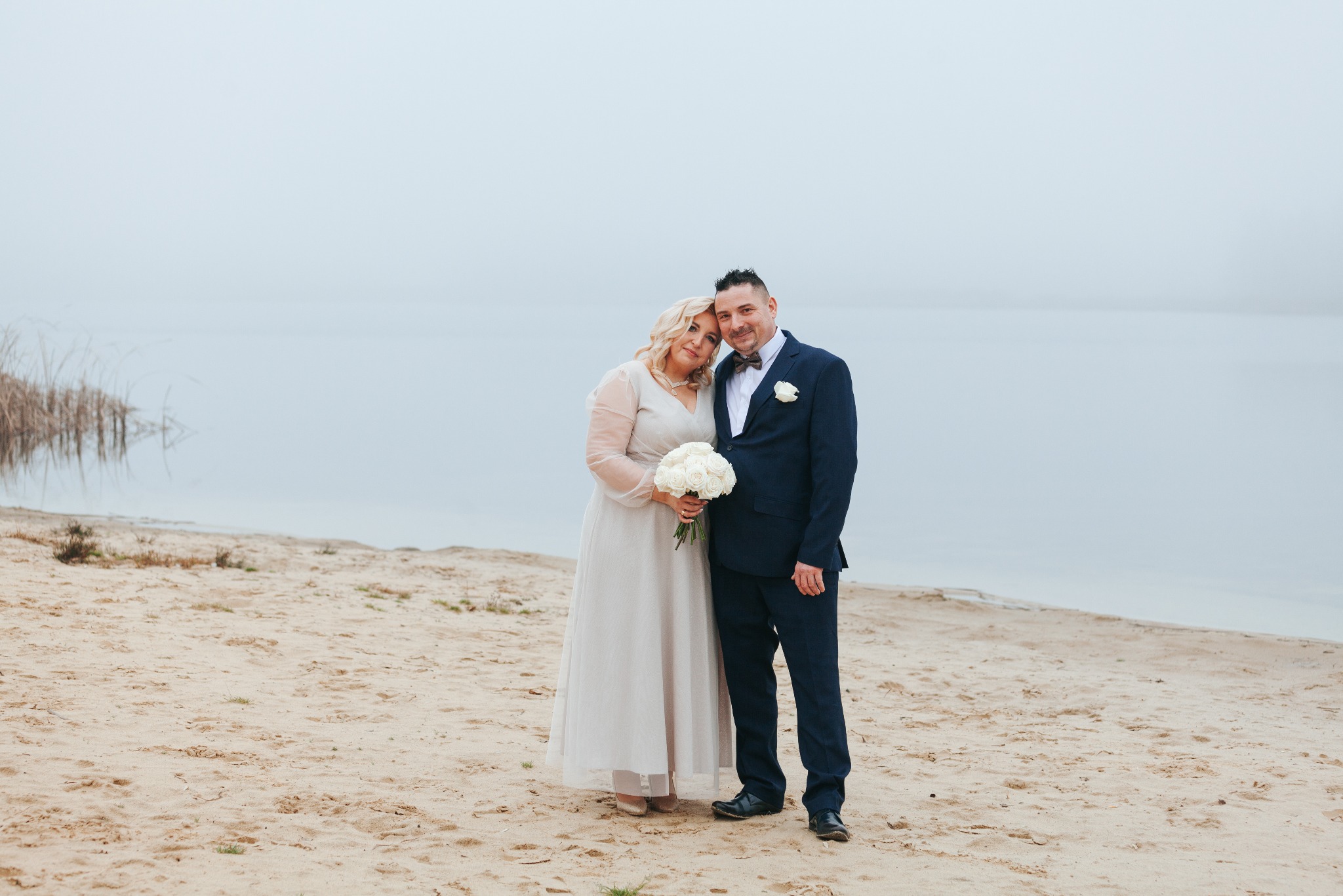 Para młoda na plaży w dniu ślubu. Panna młoda w jasnej sukni, pan młody w granatowym garniturze. W tle woda i mgła. Bukiet białych róż.