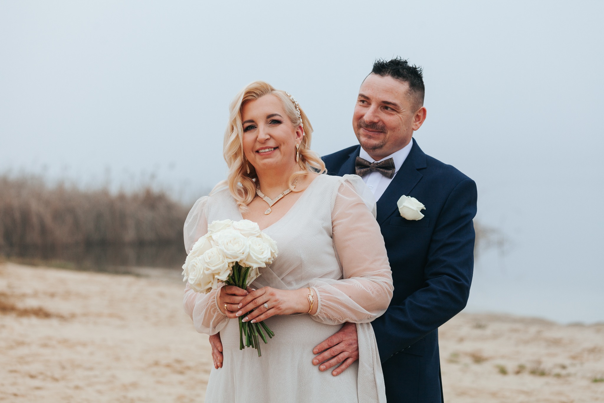 Para młoda w objęciach na plaży w pochmurny dzień. Panna młoda trzyma bukiet białych róż, a pan młody ma na sobie granatowy garnitur z białą różą w butonierce.