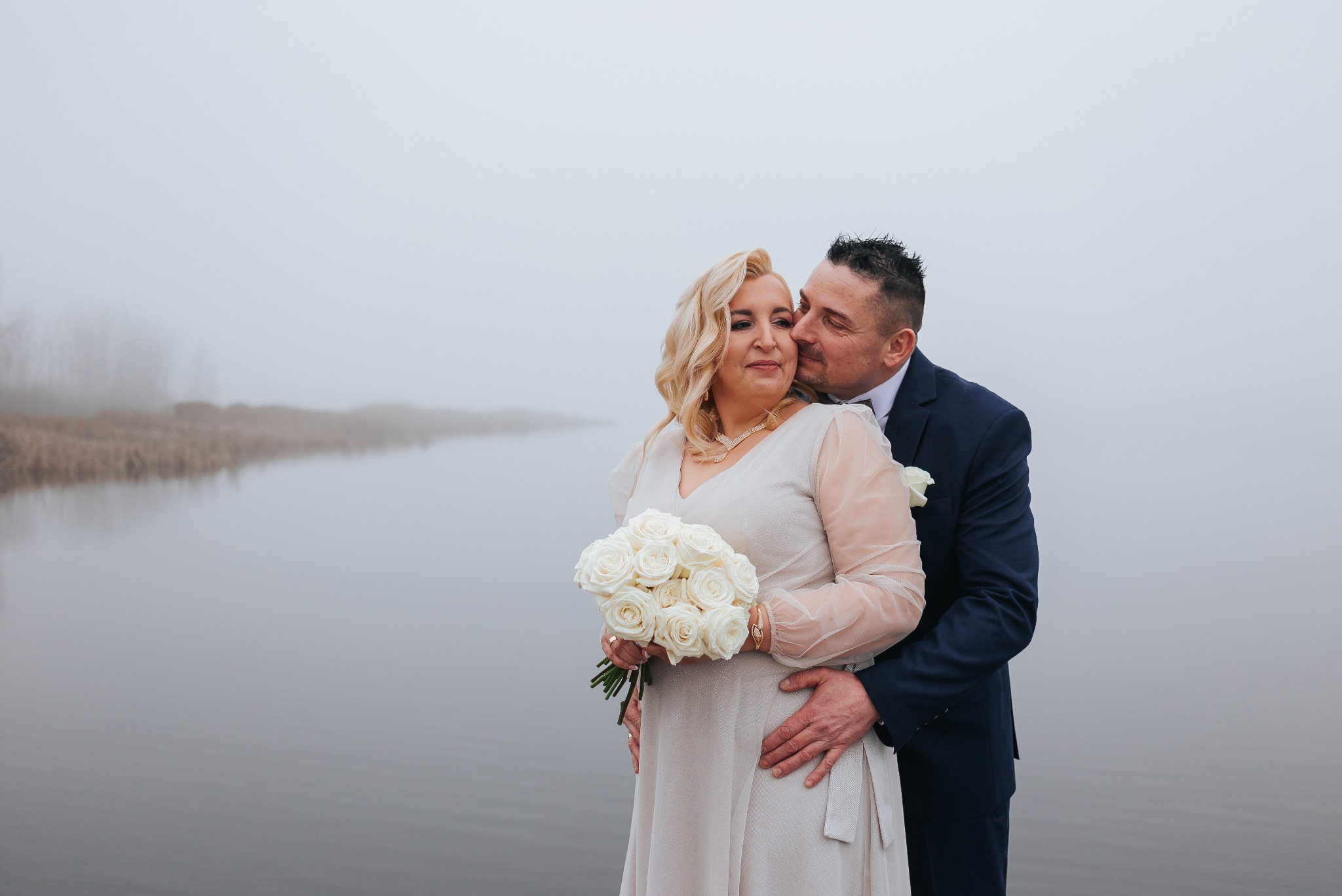 Para w strojach ślubnych, on w granatowym garniturze, ona z bukietem białych róż, w objęciach na tle mglistego krajobrazu nad wodą. Delikatne światło.