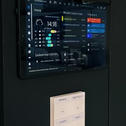 GoSmartHome Mariusz Wargocki - Tablet z interfejsem inteligentnego domu, prezentujący aktualne zużycie energii, temperaturę i stan urządzeń, zamontowany na ciemnej ścianie obok panelu sterowania z przyciskami oznaczonymi jako...