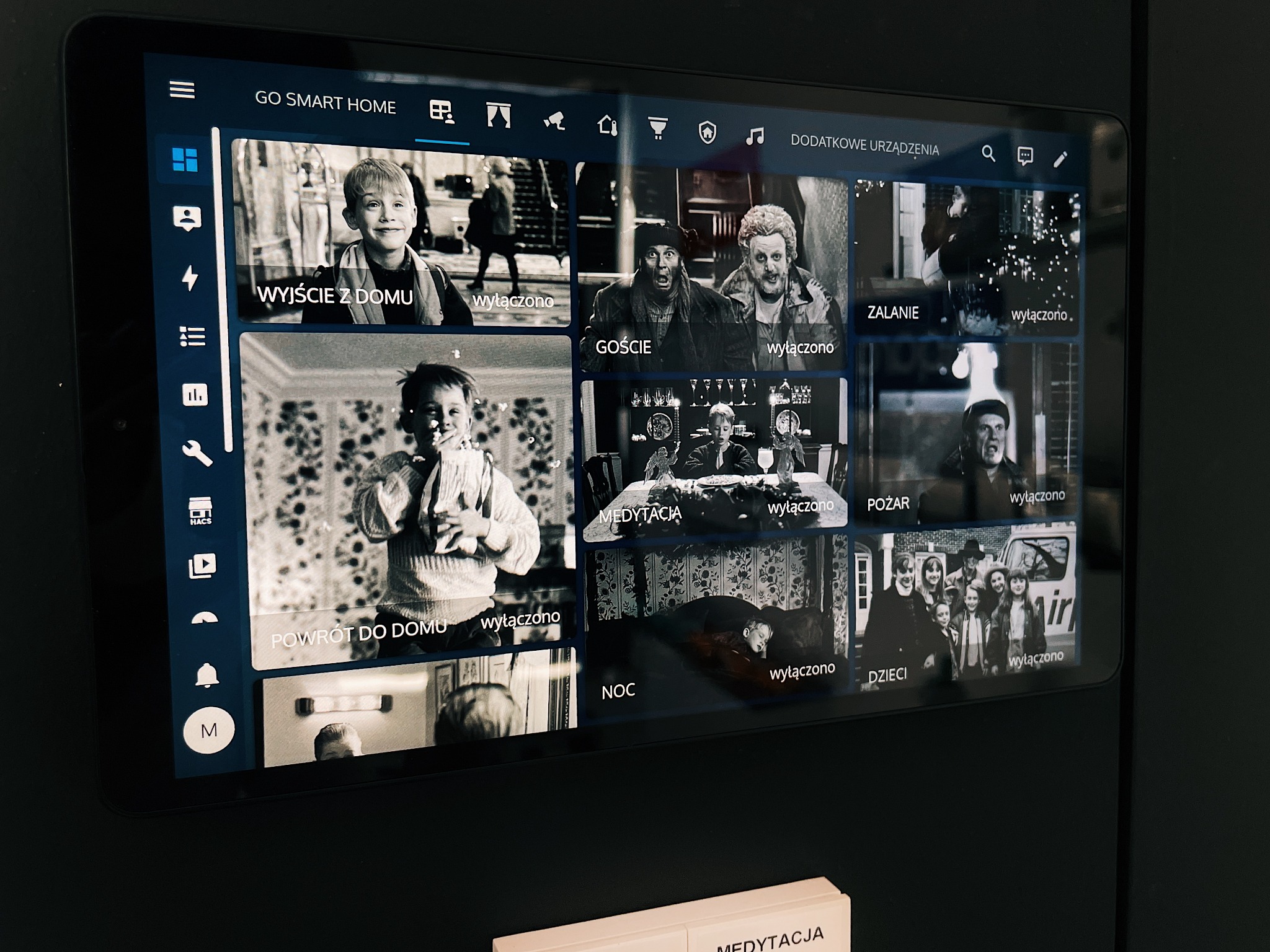 Tablet z interfejsem systemu inteligentnego domu, prezentujący czarno-białe kadry filmowe przypisane do różnych scenariuszy: wyjście z domu, goście, zalanie, powrót do domu, medytacja, pożar, noc...