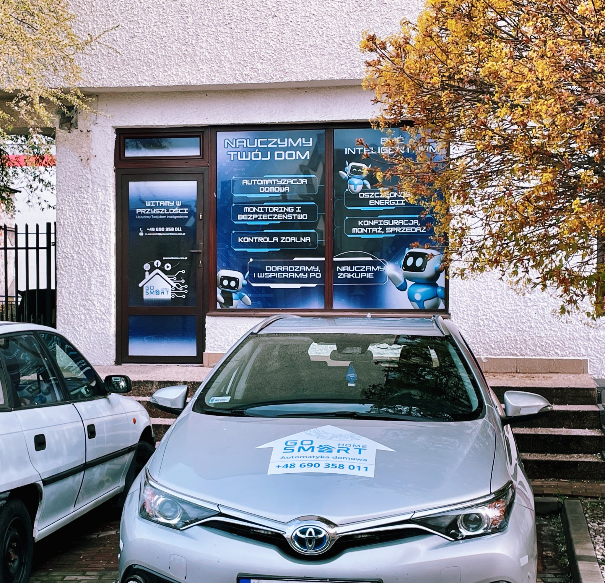 Srebrny samochód z naklejką 'Go Smart Home, Automatyka domowa' zaparkowany przed biurem firmy oferującej automatyzację domu, monitoring i bezpieczeństwo, kontrolę zdalną, konfigurację i montaż.