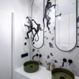 Nowoczesna łazienka z dwoma zielonymi umywalkami na białym blacie, czarną armaturą, dwoma owalnymi lustrami z dekoracją w postaci figurek małp i mozaikową ścianą w tle.