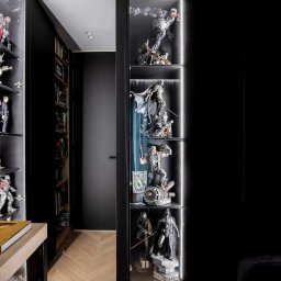 Meble Kowalski - Nowoczesna, czarna szafa wnękowa z podświetlanymi półkami, prezentująca kolekcję figurek superbohaterów i postaci z filmów science fiction, ustawiona w korytarzu z parkietem ułożonym w jodełkę.