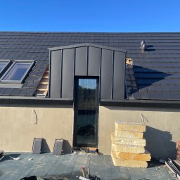 Dąb dach Paweł Dubicki - Dach z czarnej dachówki ceramicznej w trakcie prac dekarskich, z widocznym oknem dachowym i stertą izolacji. Fragment dachu bez pokrycia, narzędzia pracy w wiadrze.