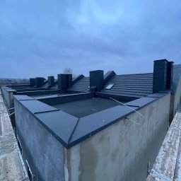 Dąb dach Paweł Dubicki - Nowoczesny dach z ciemnej dachówki ceramicznej i czarnymi kominami, widok z góry z rusztowania. Elementy blacharskie wykończone w ciemnym kolorze.