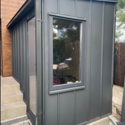 Dąb dach Paweł Dubicki - Szara, blaszana dobudówka z oknem, z widocznym systemem rynnowym Niagara, przylegająca do budynku z drewnianą elewacją. Detal architektoniczny, nowoczesny design.