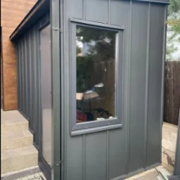 Szara, blaszana dobudówka z oknem, z widocznym systemem rynnowym Niagara, przylegająca do budynku z drewnianą elewacją. Detal architektoniczny, nowoczesny design.