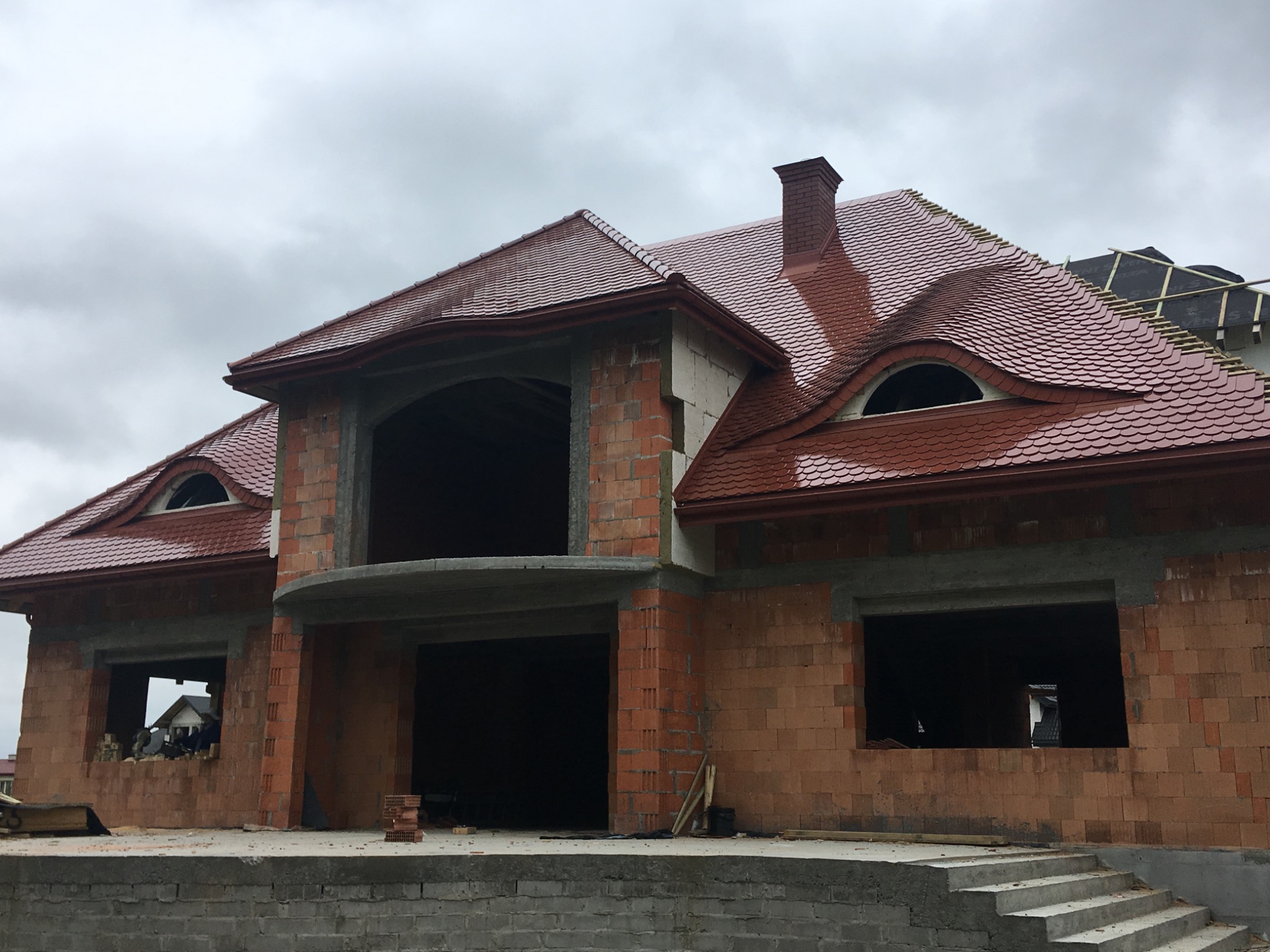 Dom w trakcie budowy z częściowo ukończonym dachem z czerwonej dachówki karpiówki, widoczne niezamknięte otwory okienne i ceglana konstrukcja ścian.