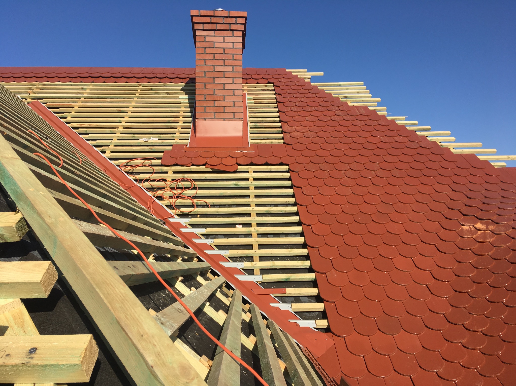 Częściowo pokryty dach z czerwonej dachówki ceramicznej w kształcie rybiej łuski, widoczna konstrukcja drewniana, komin z czerwonej cegły i niebieskie niebo w tle.