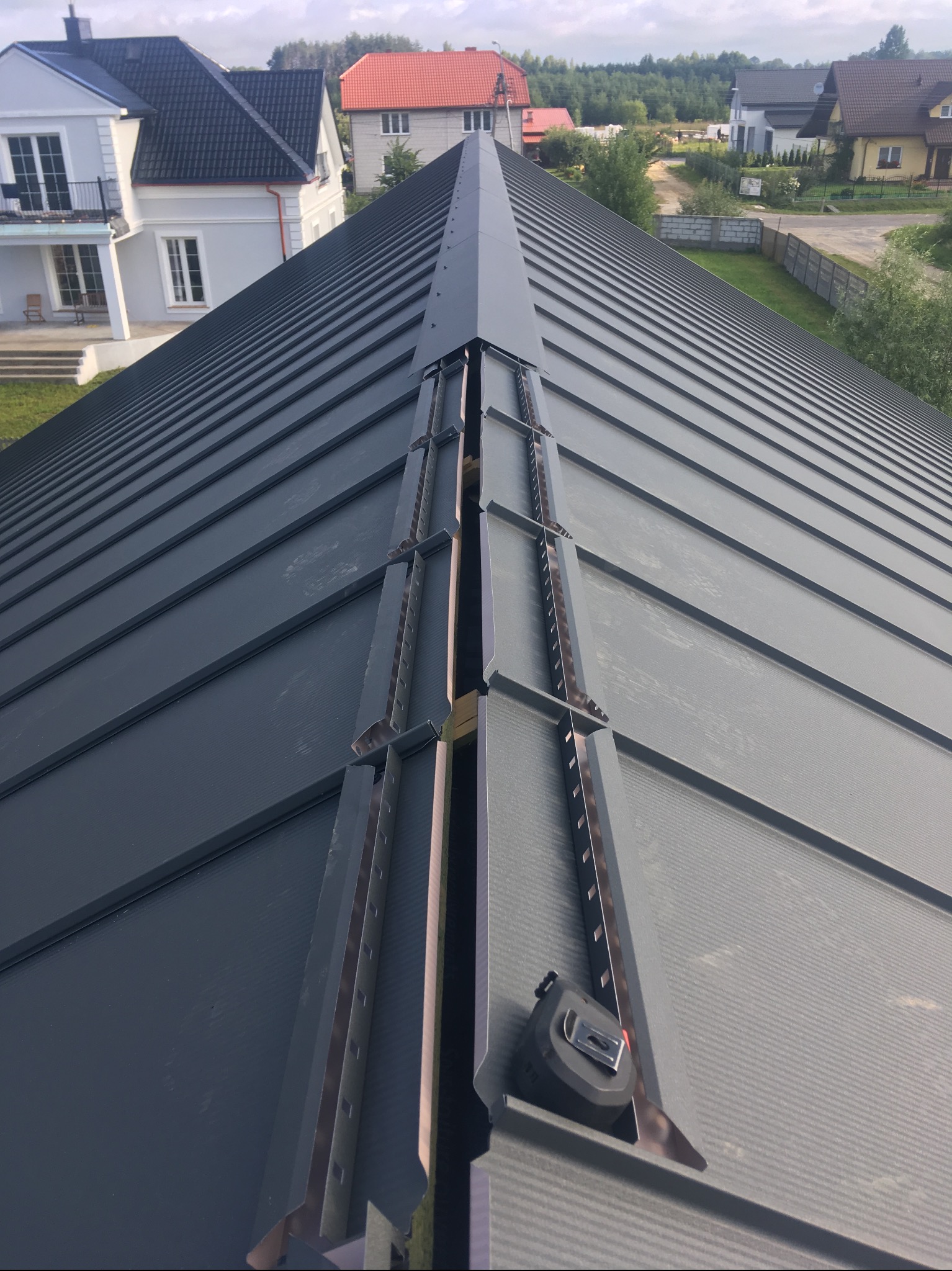 Nowoczesny, grafitowy dach z blachy na tle zabudowy jednorodzinnej, widok z góry na kalenicę z zamontowaną taśmą kalenicową i zwiniętą miarą zwisającą z krawędzi.