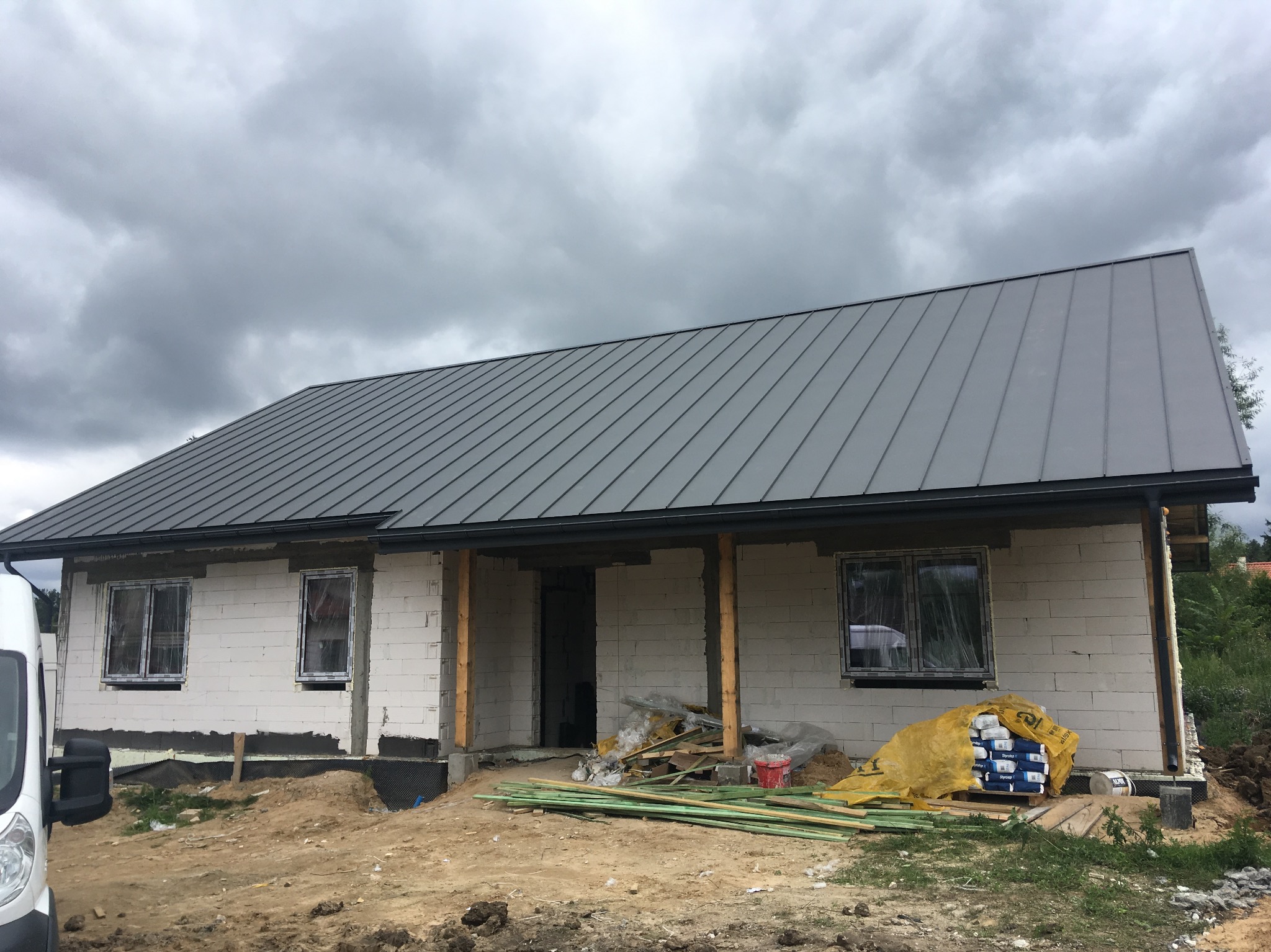 Nowy, szary dach z blachy na domu w trakcie budowy, widoczne materiały budowlane i fragment samochodu dostawczego.