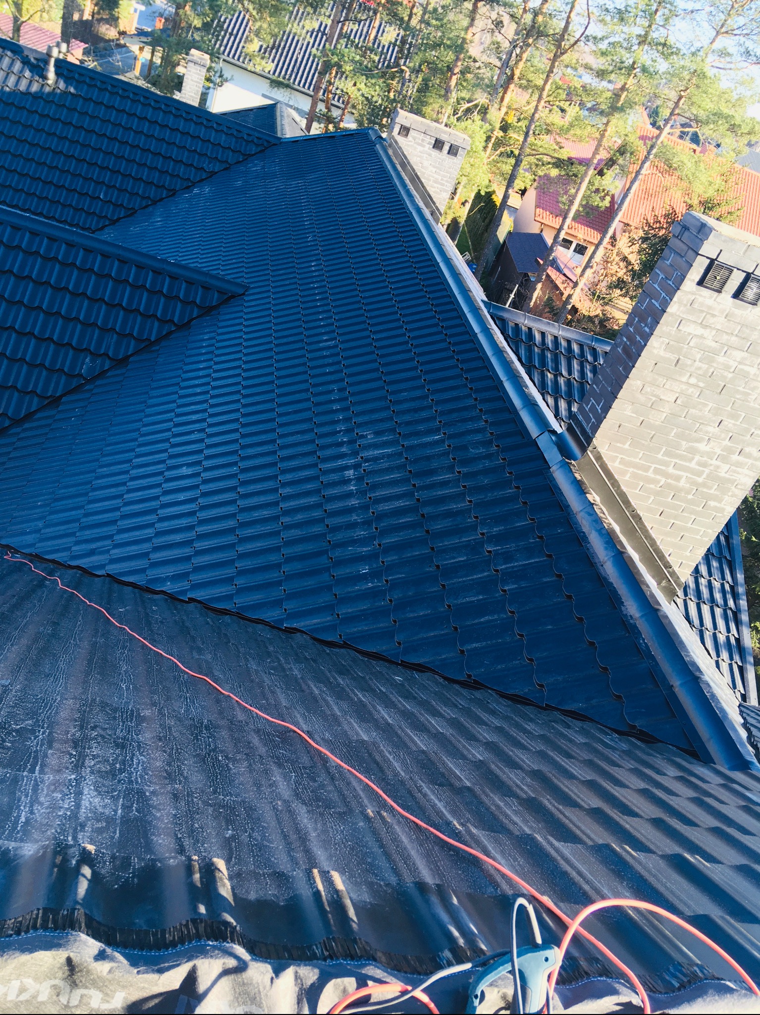 Nowy, ciemny dach z blachodachówki, widok z góry, fragment membrany dachowej z widocznymi napisami, kable i narzędzie dekarskie w dolnej części kadru.