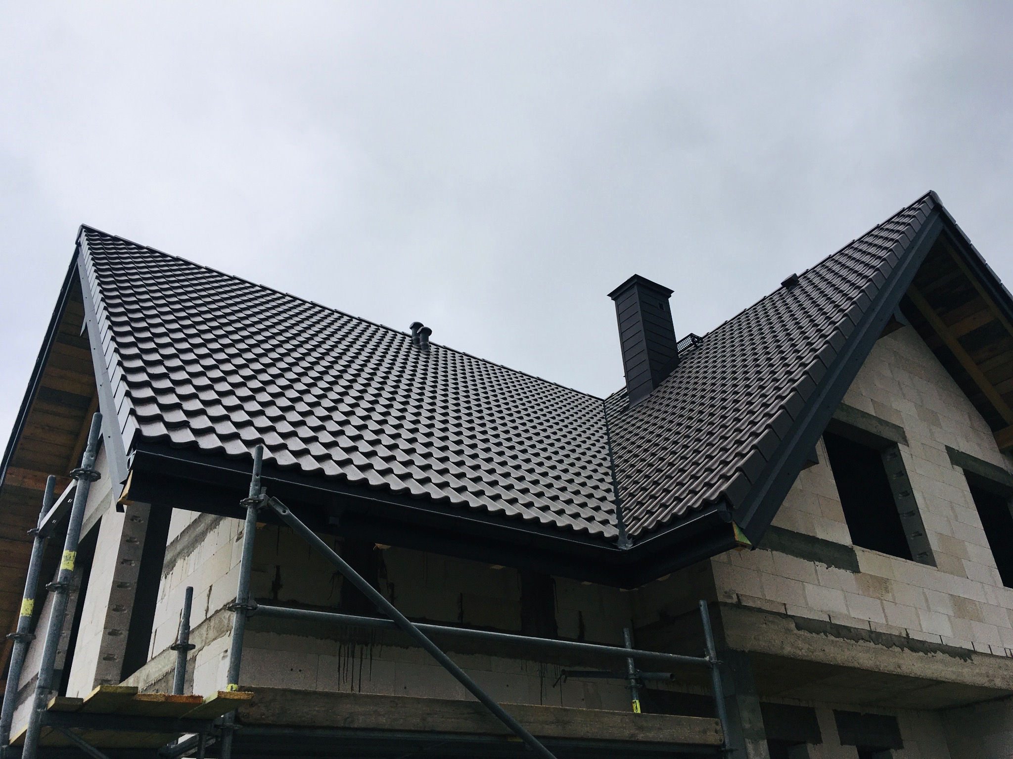 Nowy dach z czarnej blachodachówki na surowym domu w trakcie budowy, widoczne rusztowania i komin.