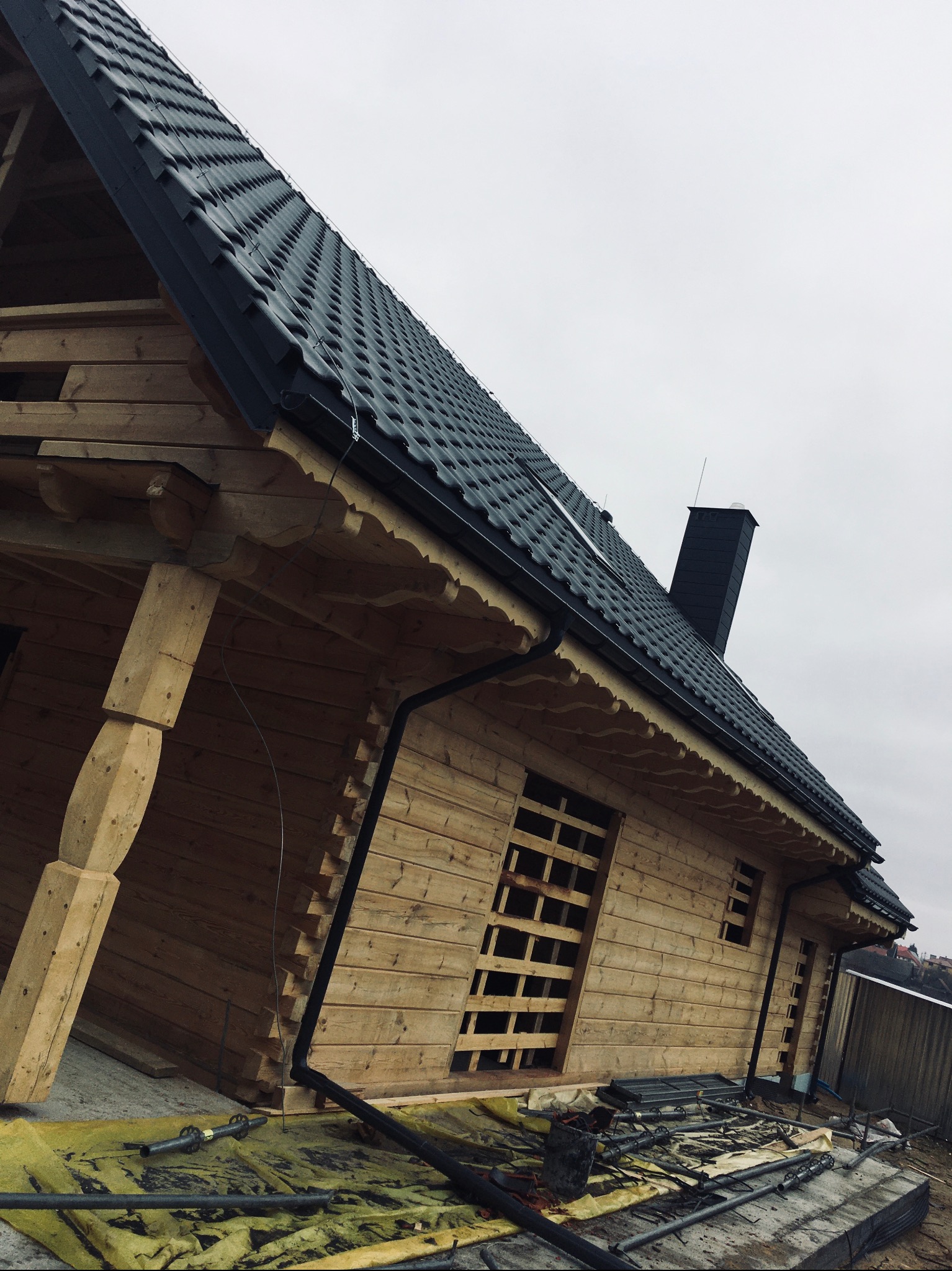 Dom z bali w trakcie budowy, widoczny dach z czarnej blachy, rynny i komin, okna zabite deskami, teren budowy z narzędziami i materiałami.