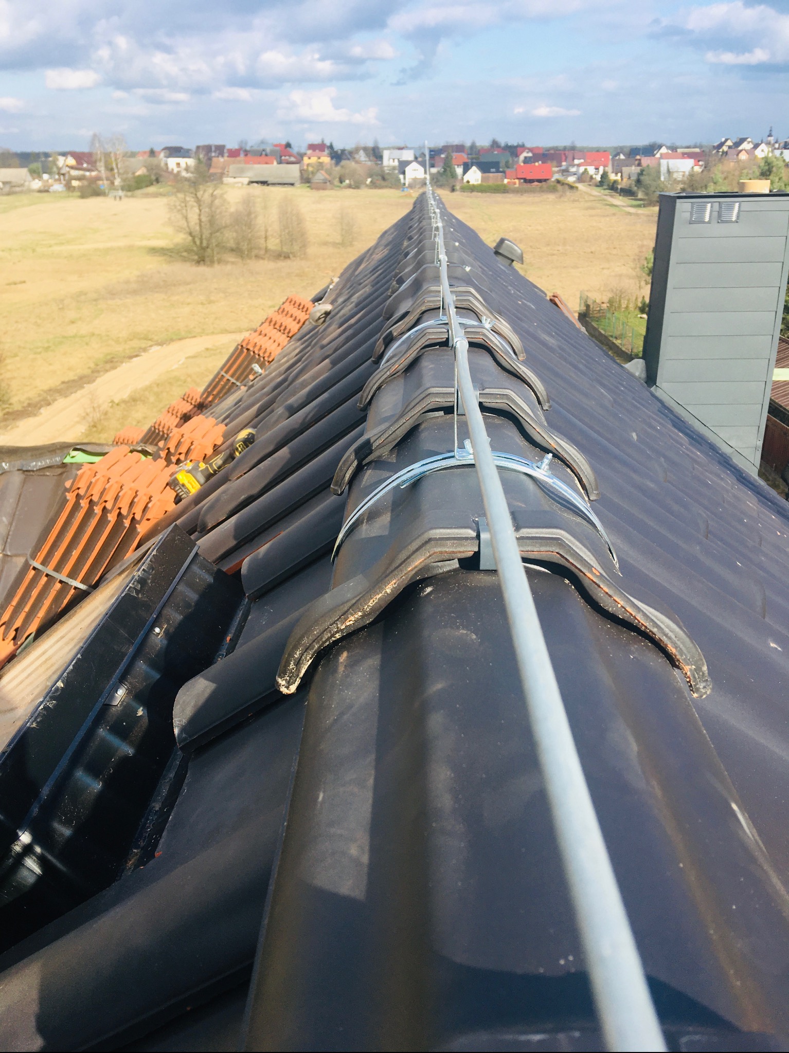 Czarny dach z ceramicznej dachówki z zamontowanym systemem odgromowym, widok z góry na tle wiejskiego krajobrazu.