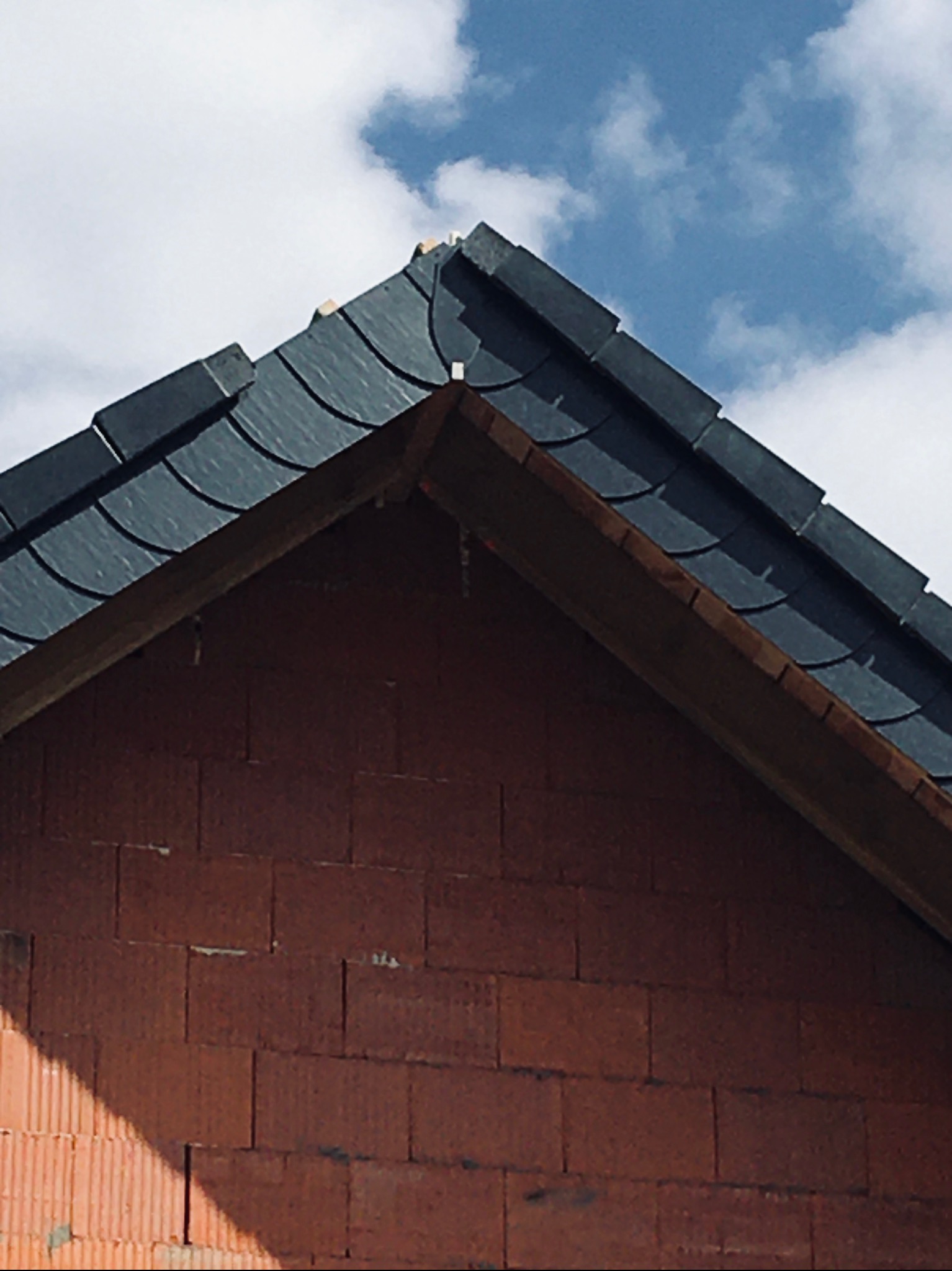 Fragment dachu krytego ciemną dachówką łuskową, widok na szczyt budynku z cegły ceramicznej, błękitne niebo z chmurami w tle.