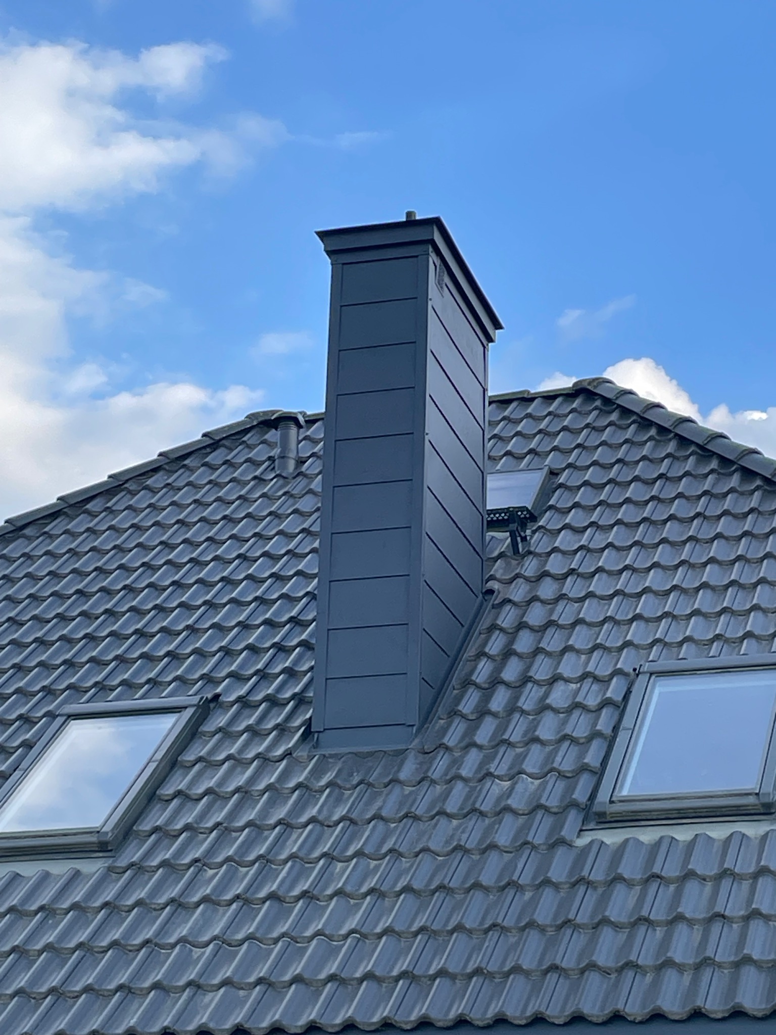 Szary dach z dachówką ceramiczną, okna dachowe i nowoczesny, metalowy komin na tle błękitnego nieba z chmurami.