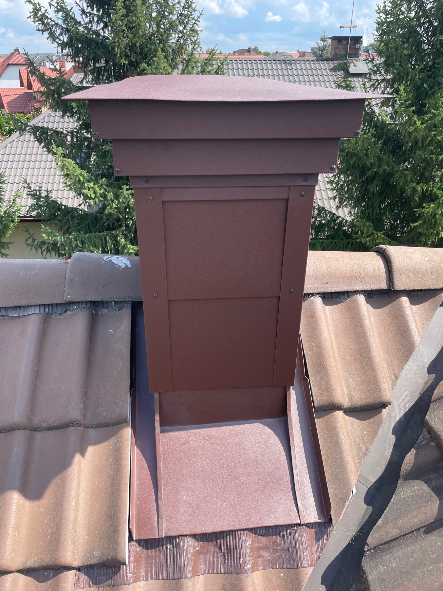 Nowy, brązowy komin z metalową obudową zamontowany na dachu z brązowej dachówki, widok z góry.