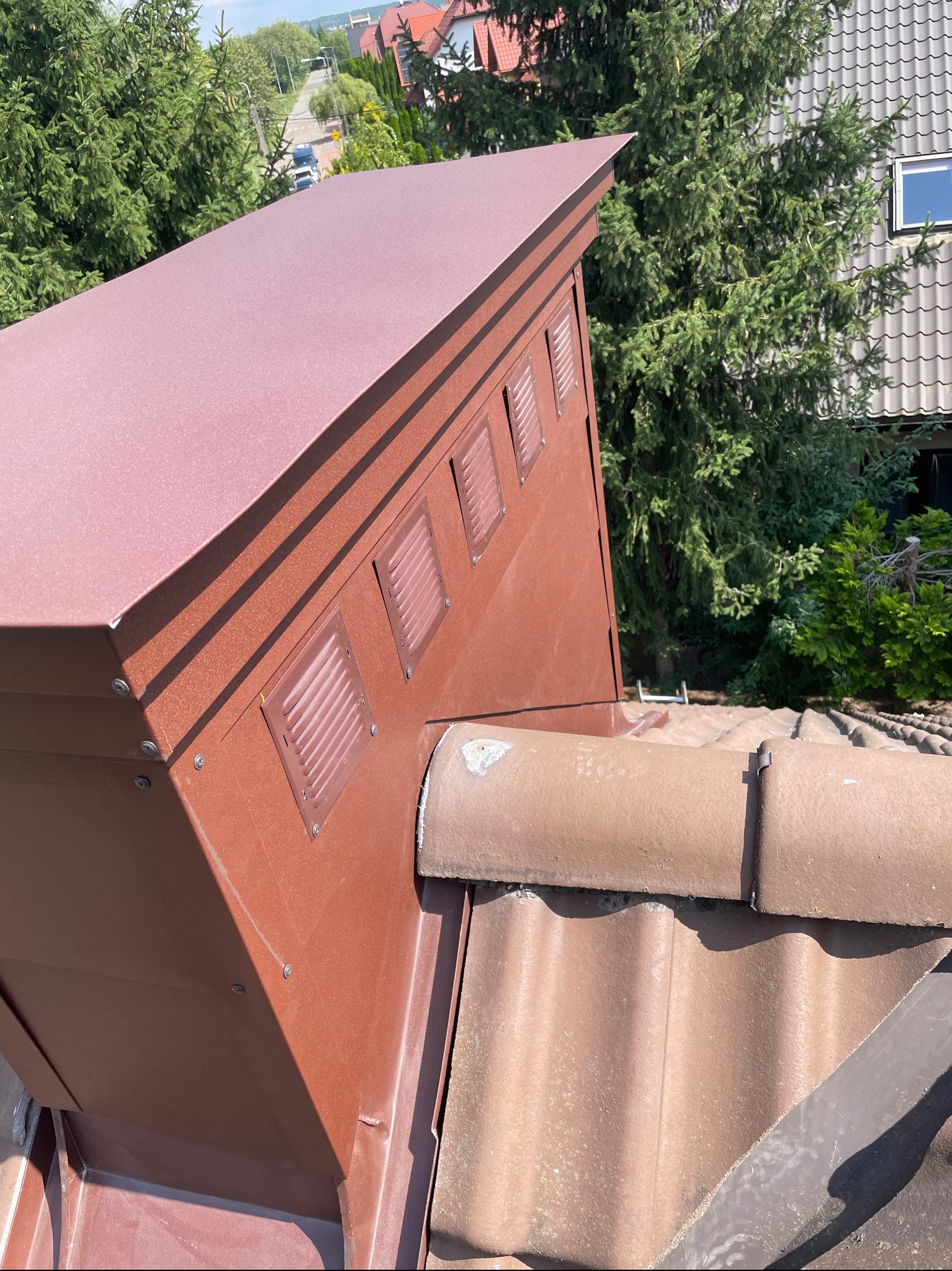 Szczegółowe ujęcie brązowej obróbki blacharskiej komina z wentylacją, zamocowanej na dachu pokrytym dachówką ceramiczną, z widokiem na zielone drzewa i zabudowania w tle.