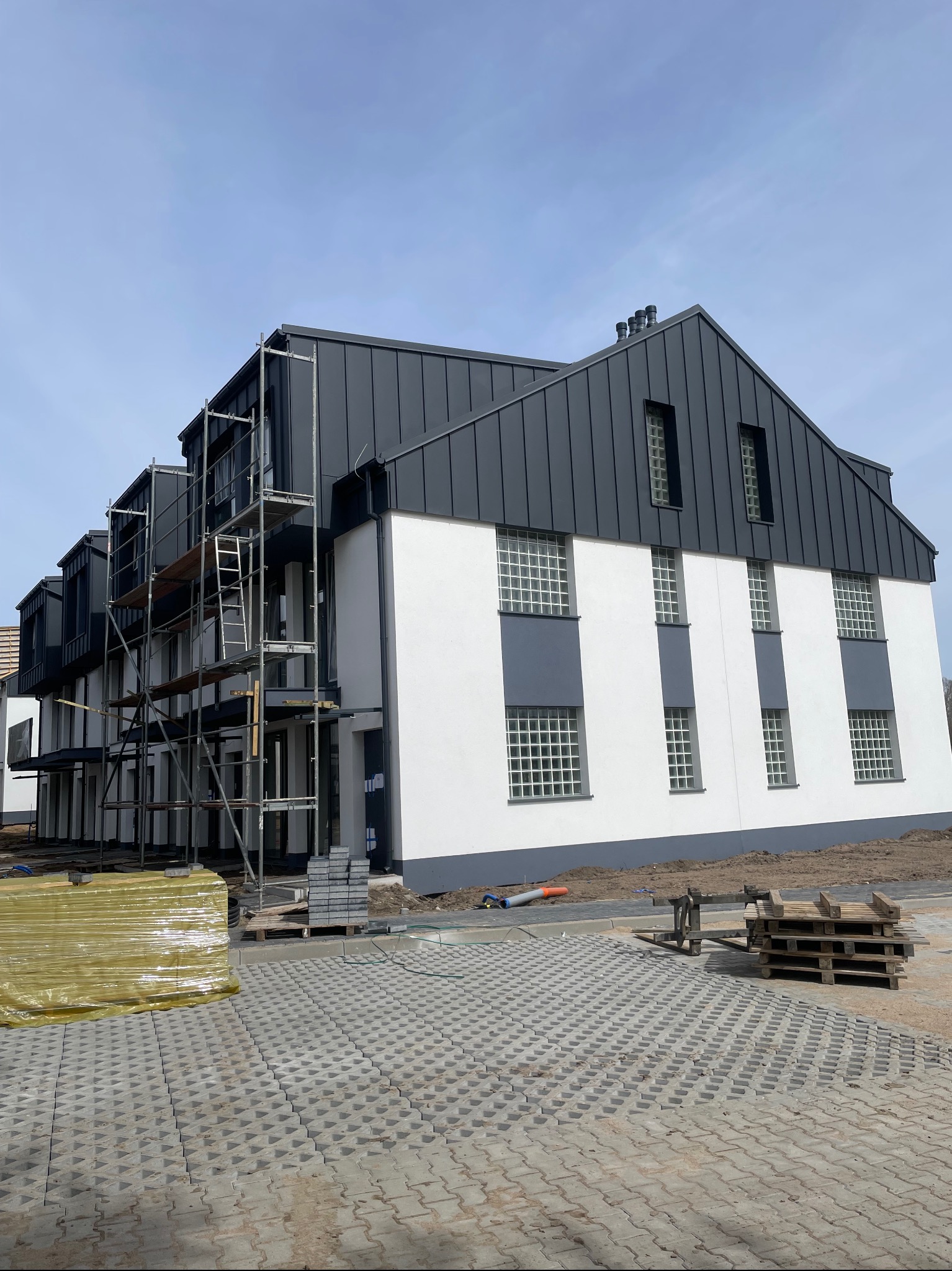 Budynek wielorodzinny z grafitowym dachem z blachy na rąbek stojący, widoczne rusztowania i palety z materiałami budowlanymi. Elementy wykończenia.