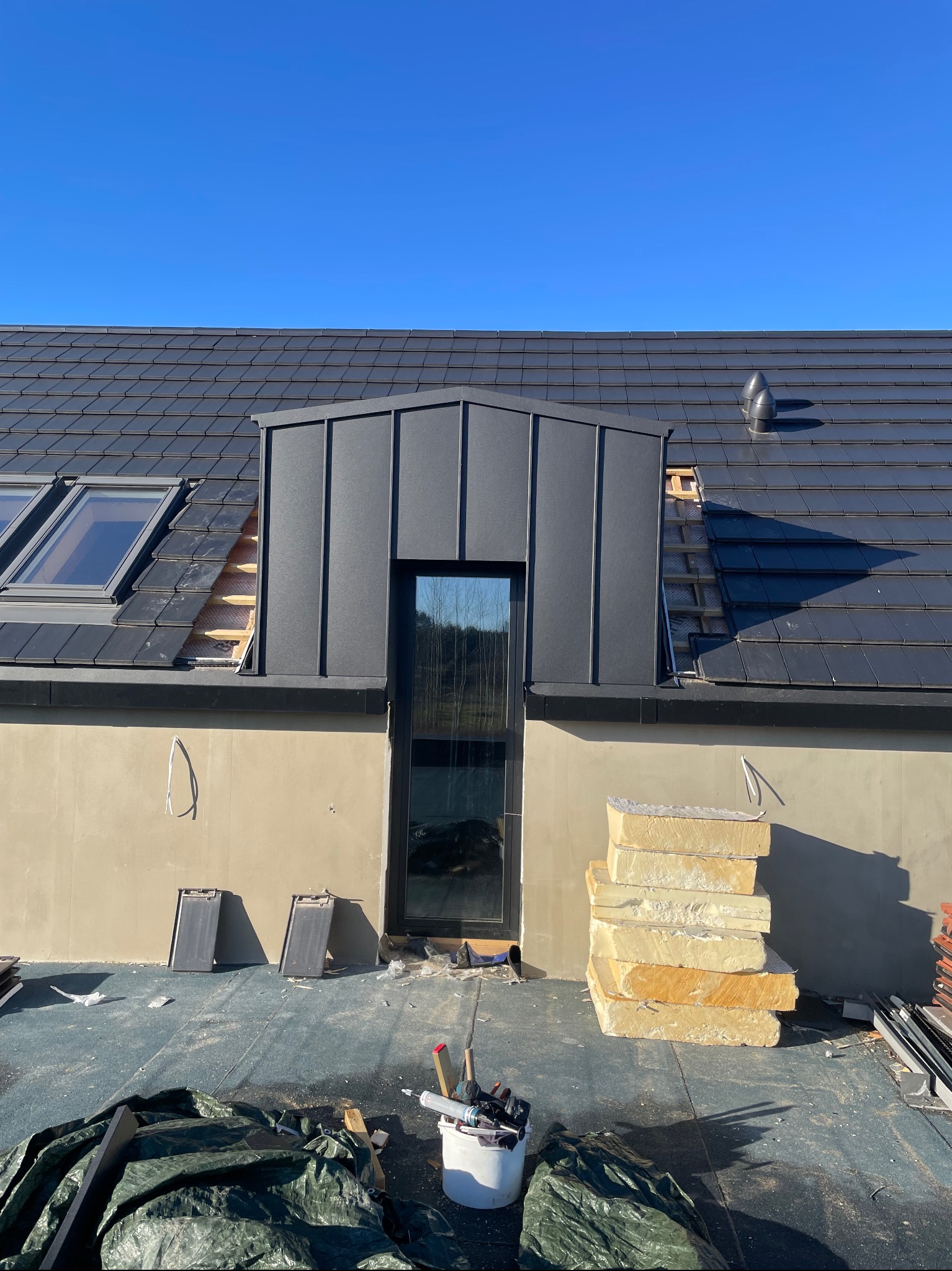 Dach z czarnej dachówki ceramicznej w trakcie prac dekarskich, z widocznym oknem dachowym i stertą izolacji. Fragment dachu bez pokrycia, narzędzia pracy w wiadrze.