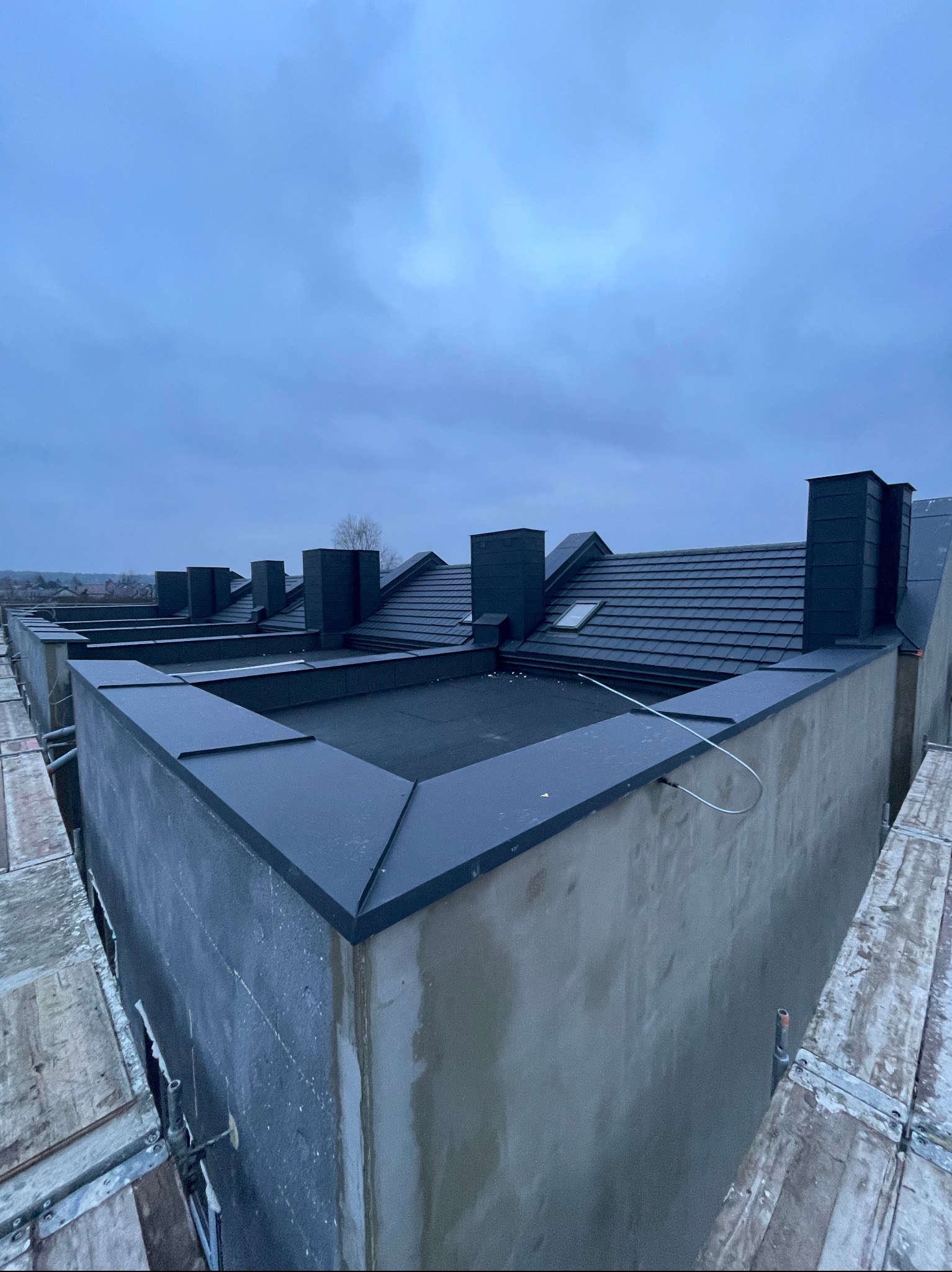 Nowoczesny dach z ciemnej dachówki ceramicznej i czarnymi kominami, widok z góry z rusztowania. Elementy blacharskie wykończone w ciemnym kolorze.