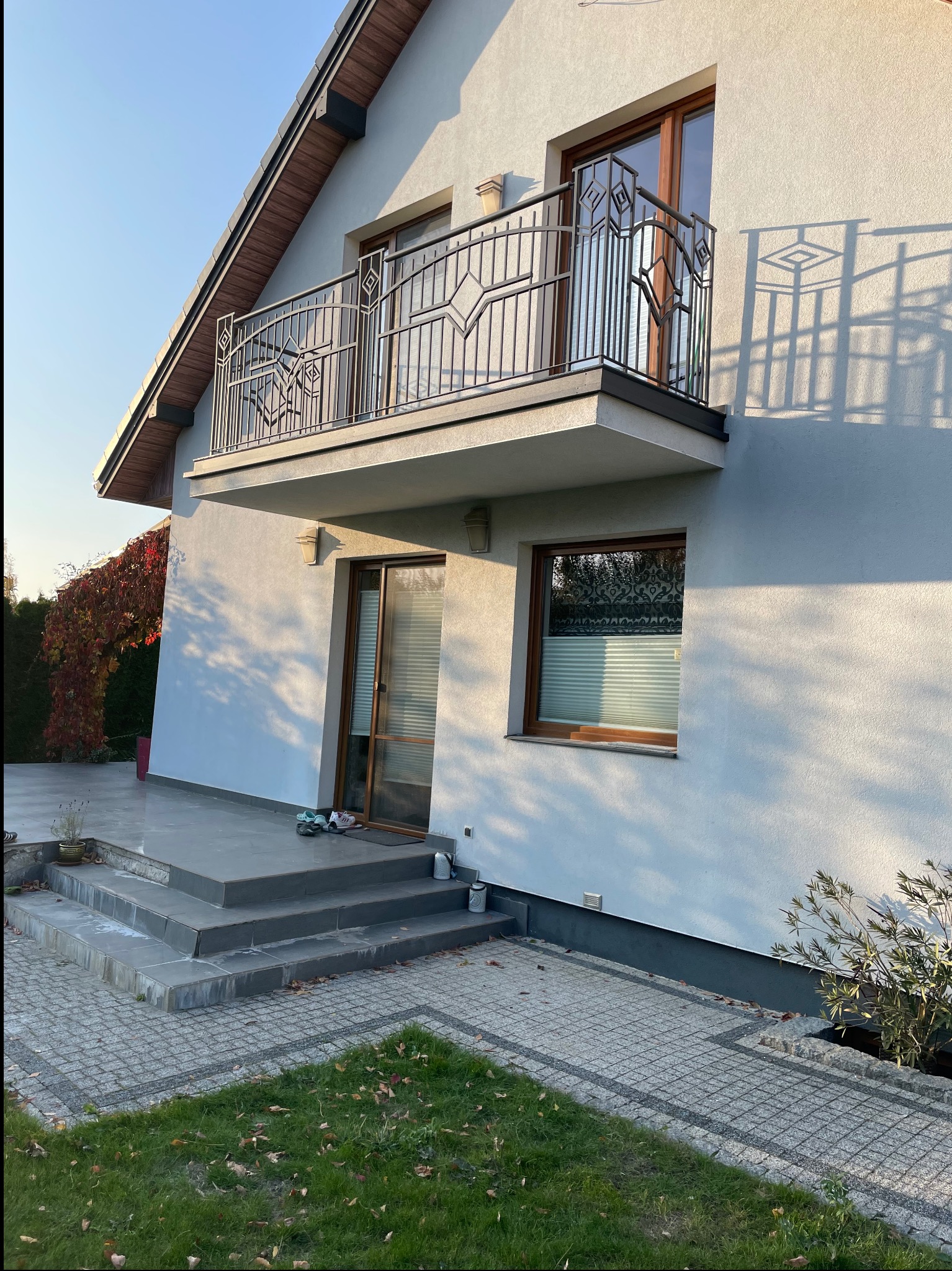 Elewacja domu z balkonem o metalowej balustradzie w geometryczne wzory. Widoczne schody wejściowe i fragment ogrodu z kostką brukową.