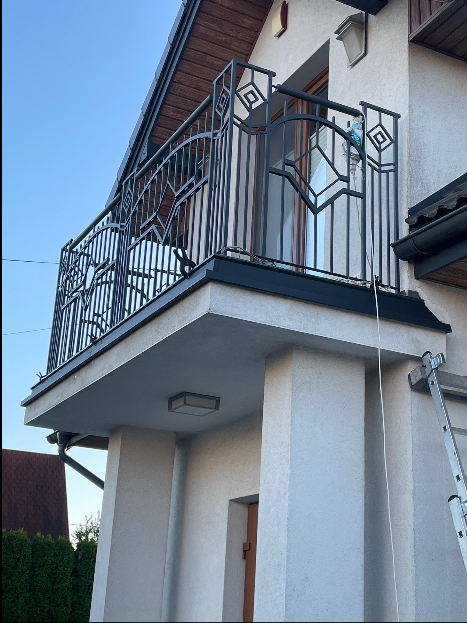 Elegancki balkon z czarną, metalową balustradą o geometrycznym wzorze,  zamontowany przy jasnym budynku z drewnianym wykończeniem dachu.  Widoczna drabina.