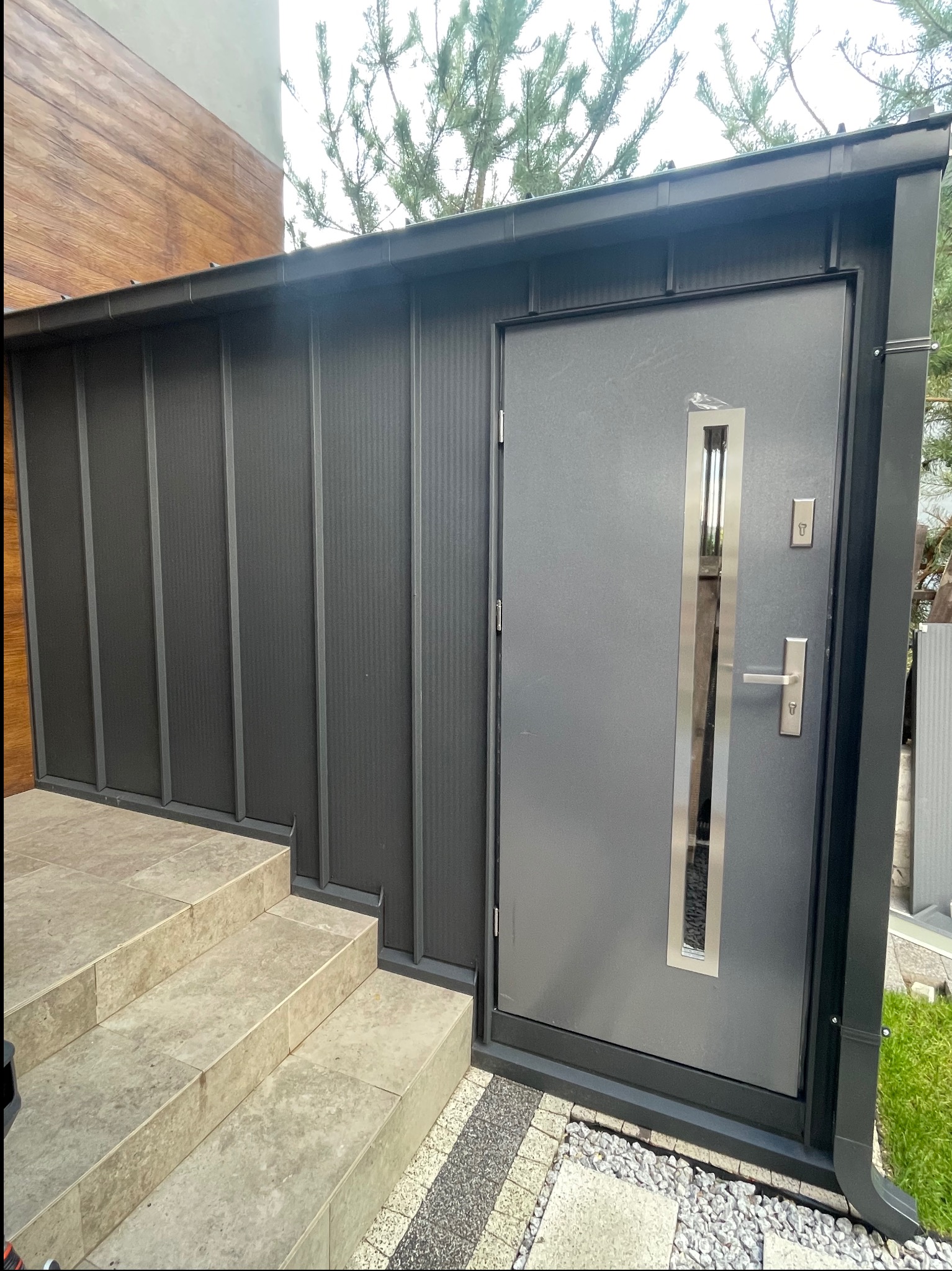 Szara, nowoczesna elewacja domu z metalowym panelem i drzwiami wejściowymi z ozdobnym, pionowym przeszkleniem oraz rynną w odcieniu grafitu.