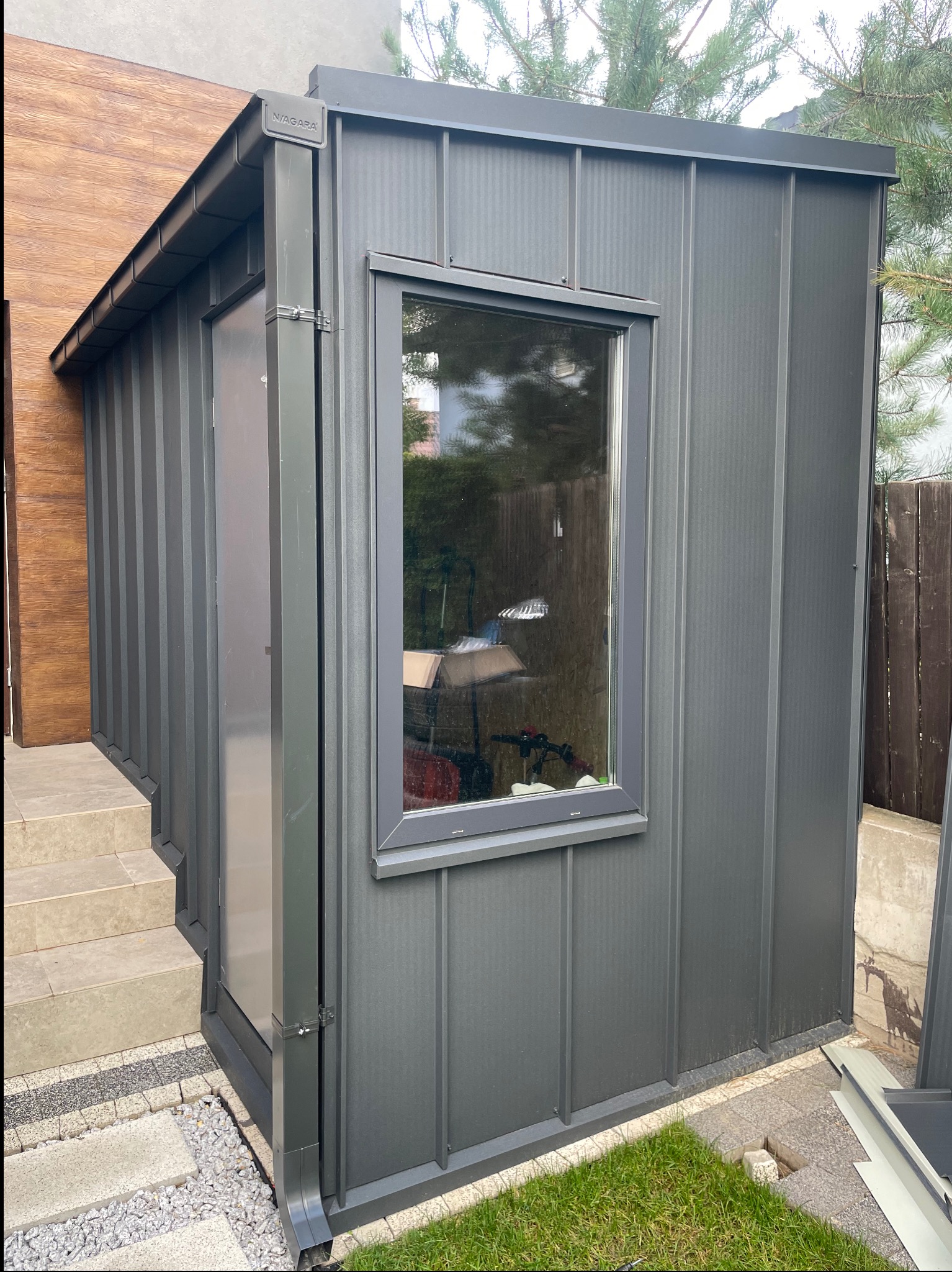 Szara, blaszana dobudówka z oknem, z widocznym systemem rynnowym Niagara, przylegająca do budynku z drewnianą elewacją. Detal architektoniczny, nowoczesny design.