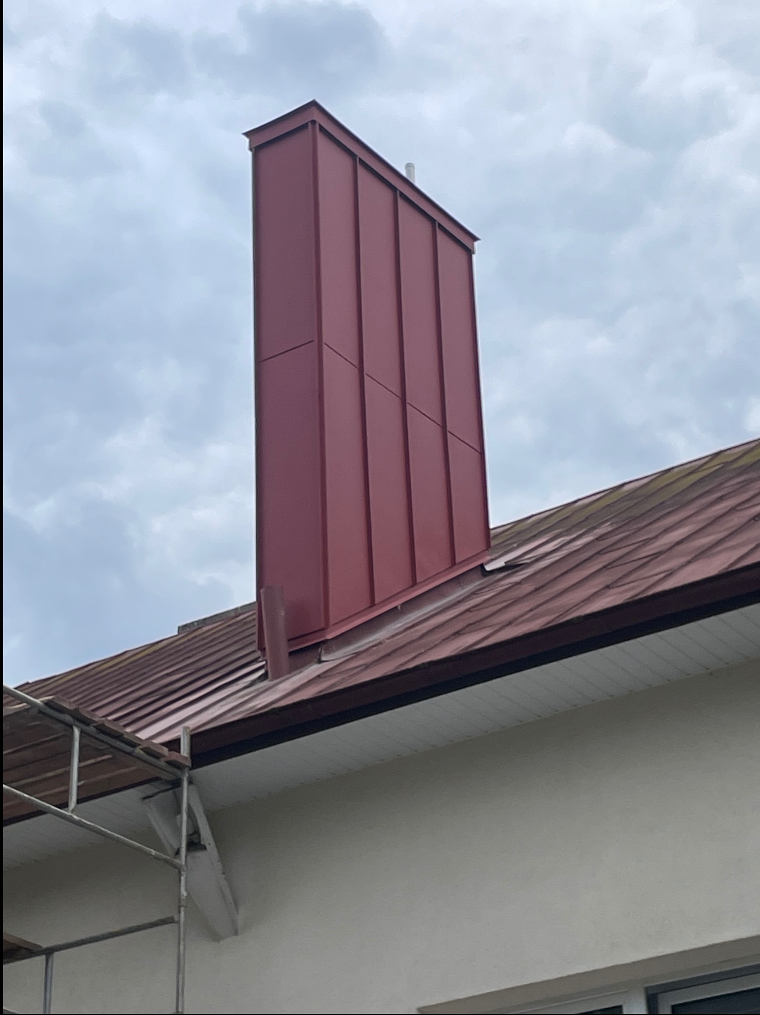 Nowoczesny, bordowy komin z blachy na dachu z dachówki ceramicznej. Widoczne fragmenty rusztowania i orynnowania. Pochmurne niebo w tle.