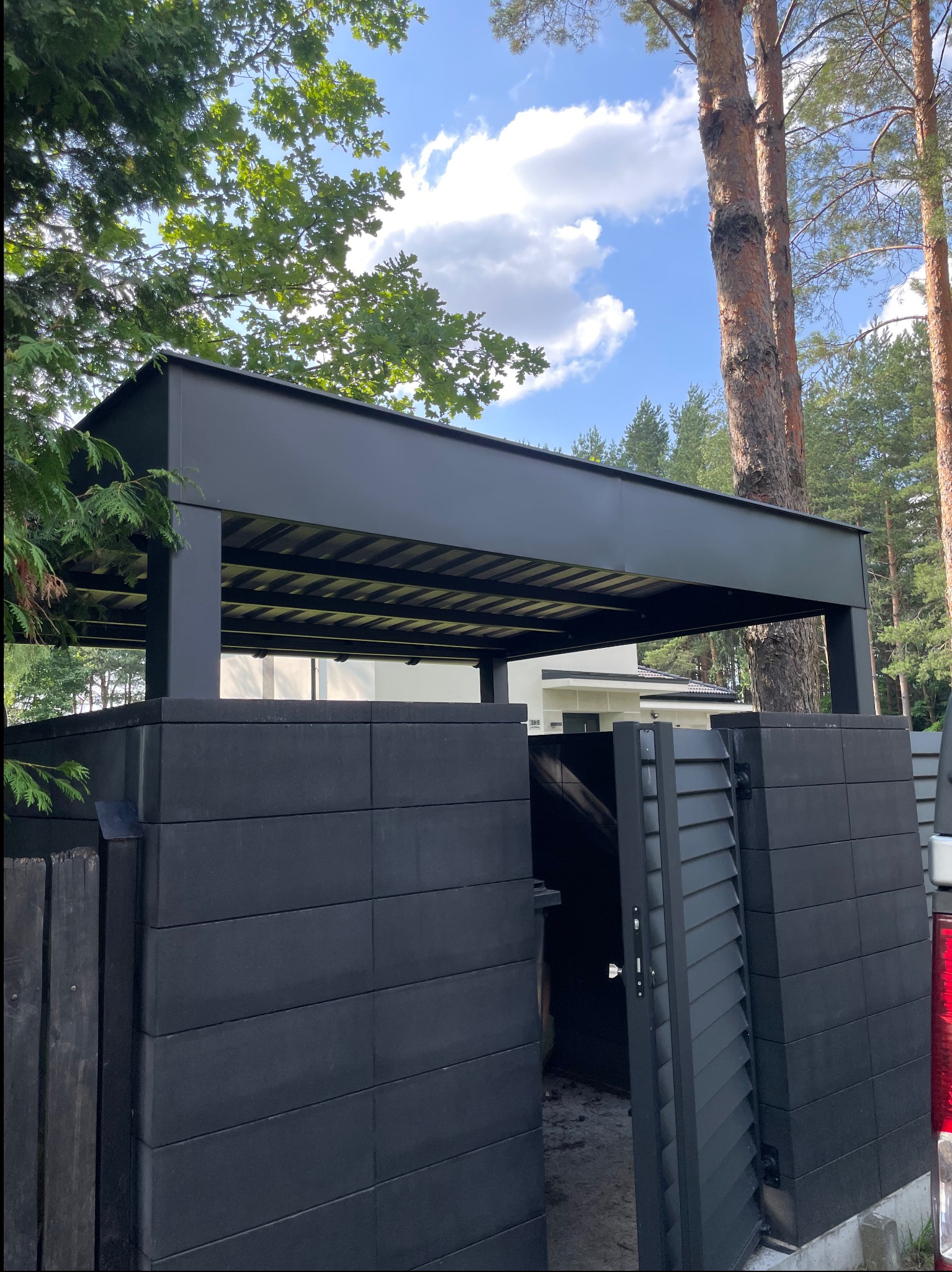 Nowoczesna, czarna konstrukcja zadaszenia nad wejściem, z elementami blacharskimi, na tle zieleni drzew i błękitnego nieba. Widoczne detale wykończenia i solidna konstrukcja.