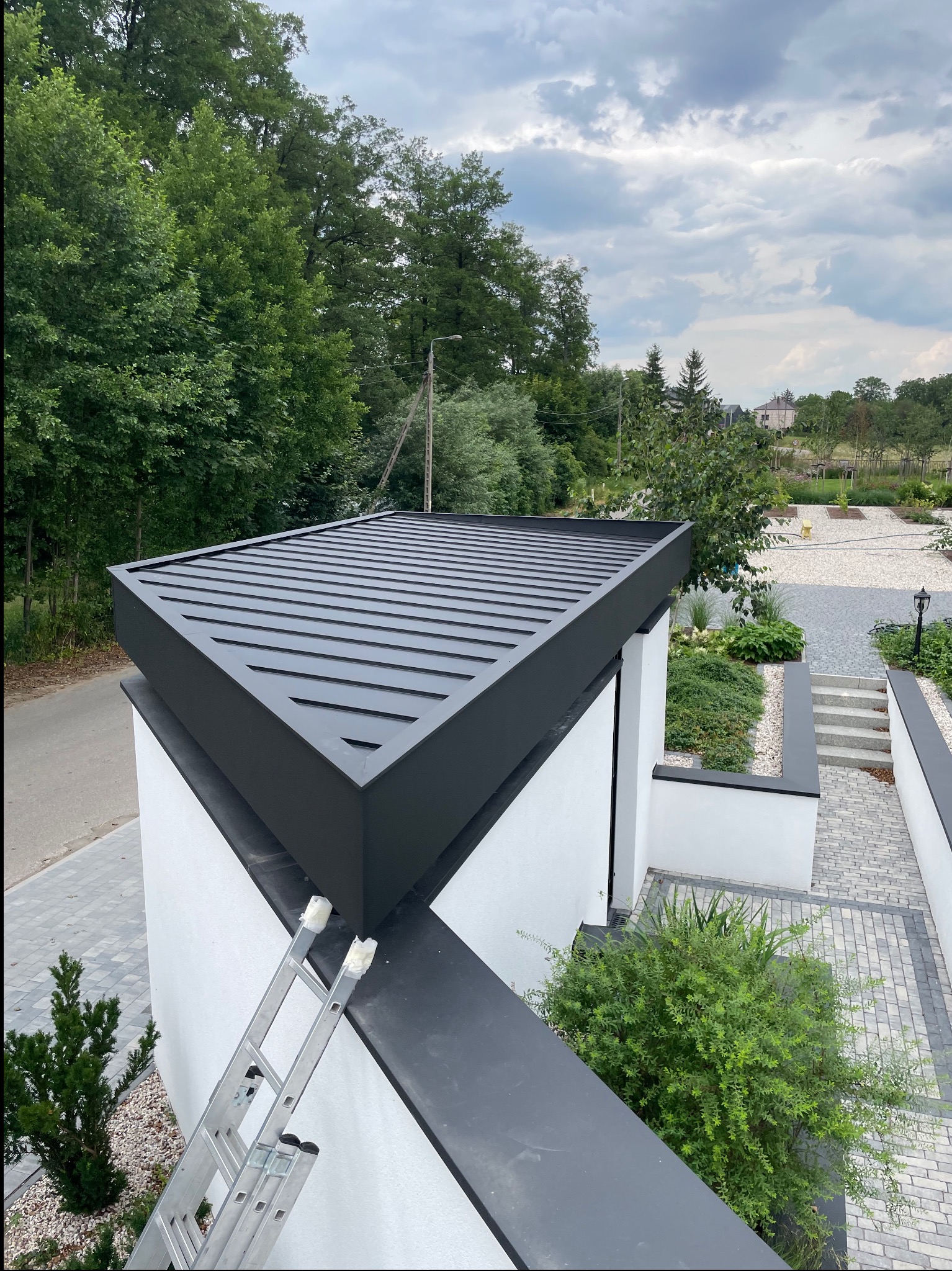 Nowoczesny dach z blachy trapezowej w kolorze antracytowym, widok z góry, Białystok. Minimalistyczny design, drabina oparta o ścianę, otoczenie zieleni.