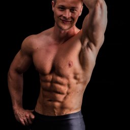 trener lublin - Uśmiechnięty, muskularny mężczyzna z ręką na głowie, prezentujący wyrzeźbiony brzuch na czarnym tle.