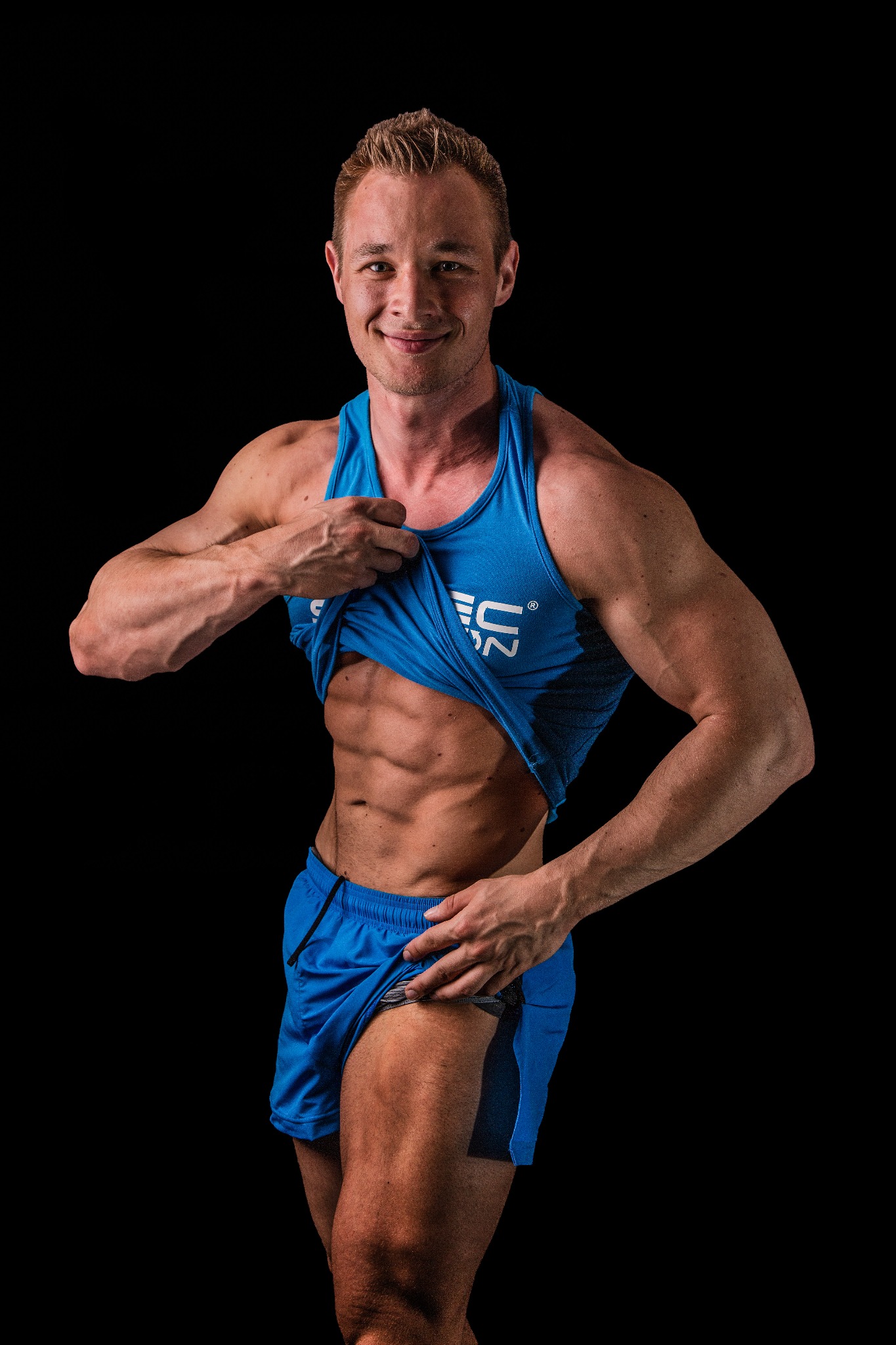 Mężczyzna w niebieskim stroju sportowym, unoszący koszulkę, prezentuje wyrzeźbiony brzuch na czarnym tle.