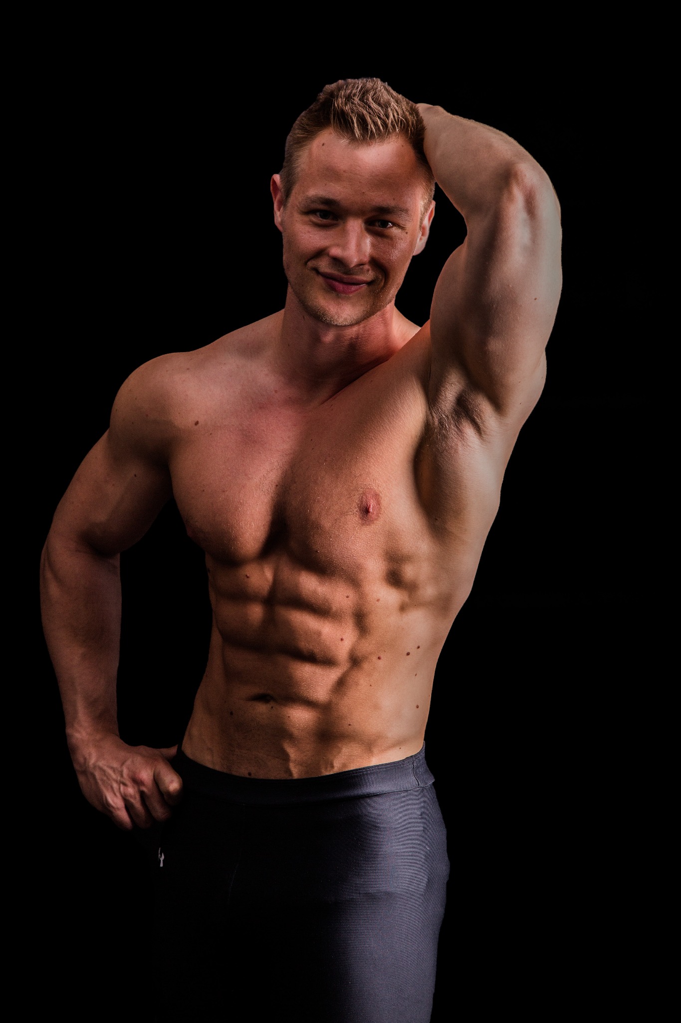 Uśmiechnięty, muskularny mężczyzna z ręką na głowie, prezentujący wyrzeźbiony brzuch na czarnym tle.