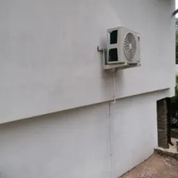 Zewnętrzna jednostka klimatyzacji marki Gree zamontowana na wsporniku na szarej, chropowatej ścianie budynku, z widocznymi rurami odprowadzającymi skropliny.