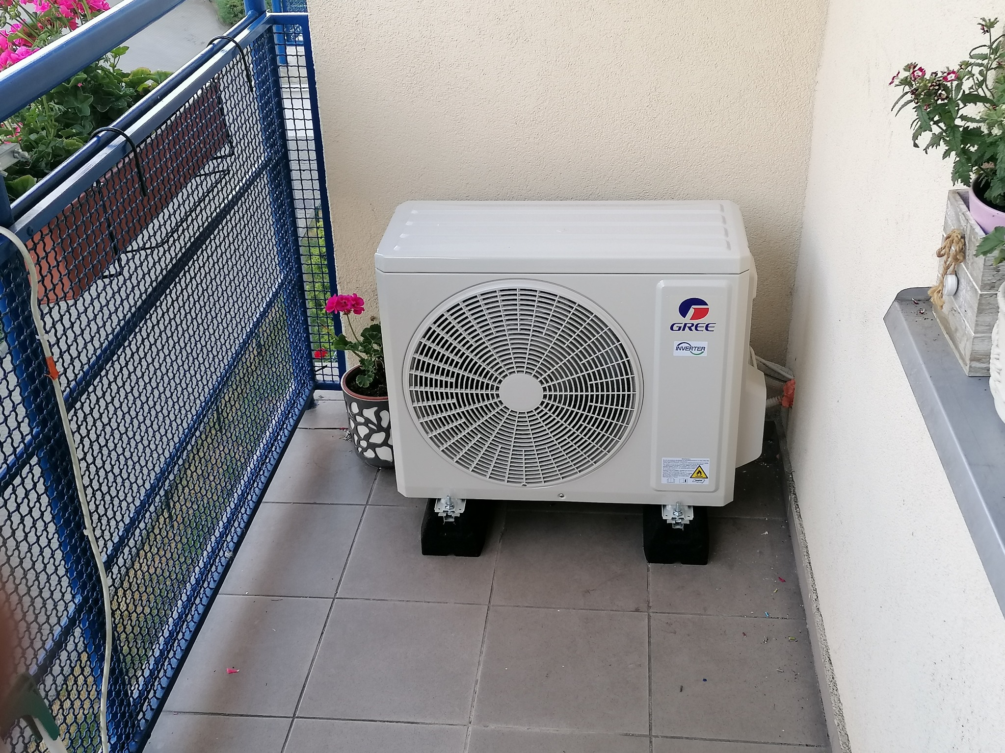 Zewnętrzna jednostka klimatyzatora marki Gree Inverter, umieszczona na balkonie, na czarnych podkładkach antywibracyjnych, z widocznymi śrubami mocującymi.