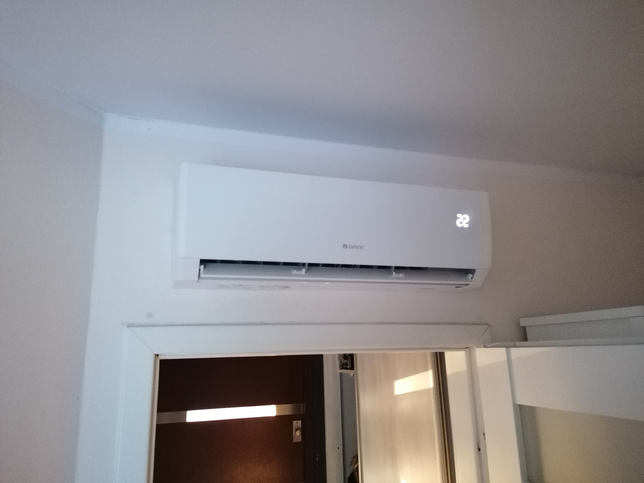 Biała klimatyzacja marki Gree zamontowana nad futryną drzwi, wyświetlająca temperaturę 22 stopnie Celsjusza.