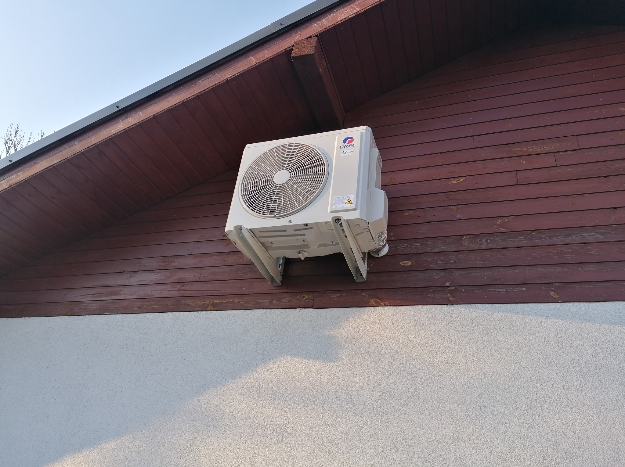Zewnętrzna jednostka klimatyzacji marki Gree zamontowana na drewnianej ścianie domu, widoczne mocowania i fragment białej elewacji poniżej.
