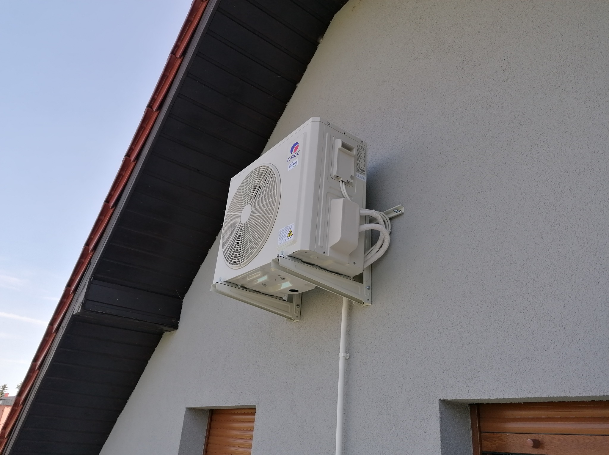 Zewnętrzna jednostka klimatyzatora marki Gree zamontowana na elewacji budynku z szarego tynku strukturalnego, widoczna część dachu z ciemną podbitką i czerwoną dachówką, przewody odprowadzające...