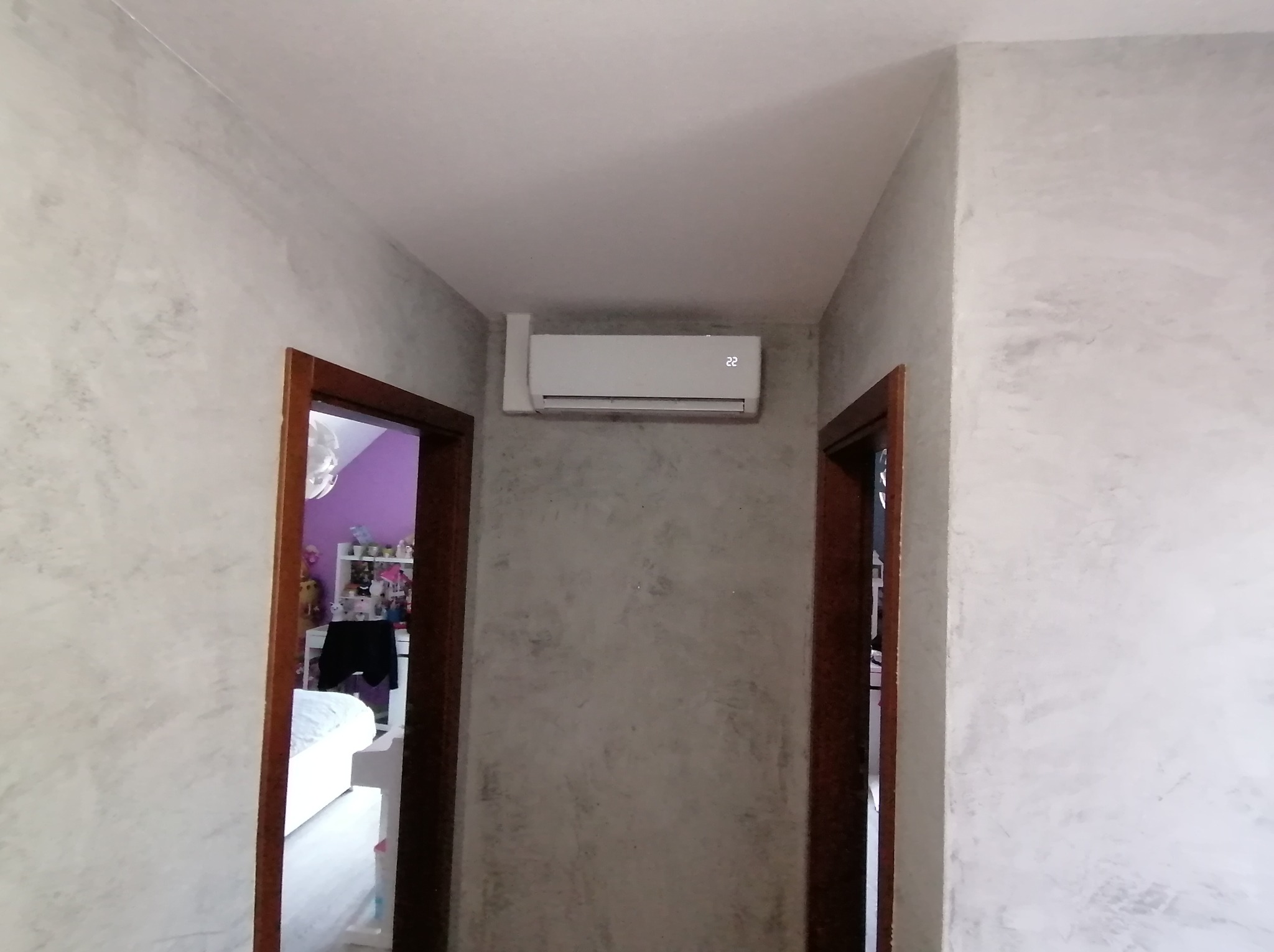 Biały klimatyzator ścienny z wyświetlaczem '22' zamontowany w korytarzu z dekoracyjną szarą ścianą, widok na pokój dziecięcy przez otwarte drzwi po lewej stronie.