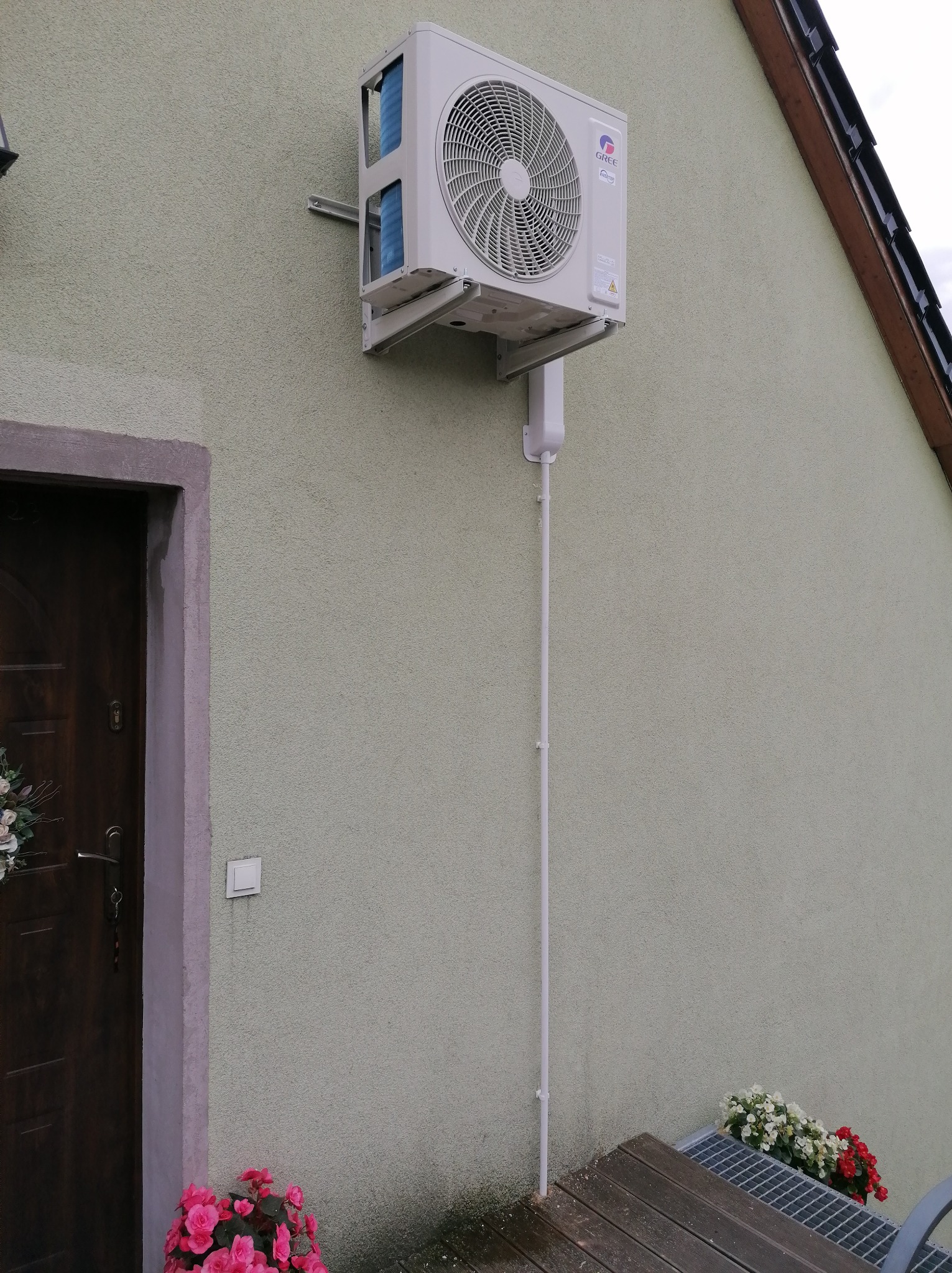 Zewnętrzna jednostka klimatyzacji firmy Gree zamontowana na elewacji domu, z widoczną rurą odprowadzającą skropliny, obok drzwi wejściowych z dekoracją kwiatową i kwiatami w doniczkach na podeście.